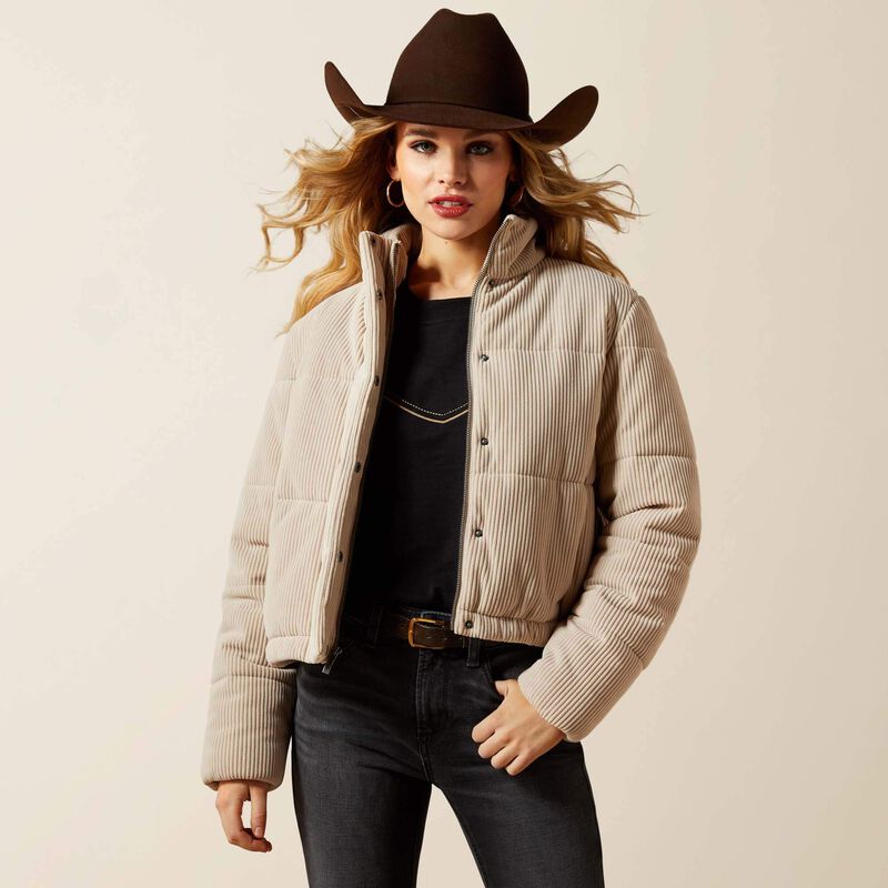 Woman wearing a beige puffer jacket, black top, dark jeans, and a brown cowboy hat on a beige background