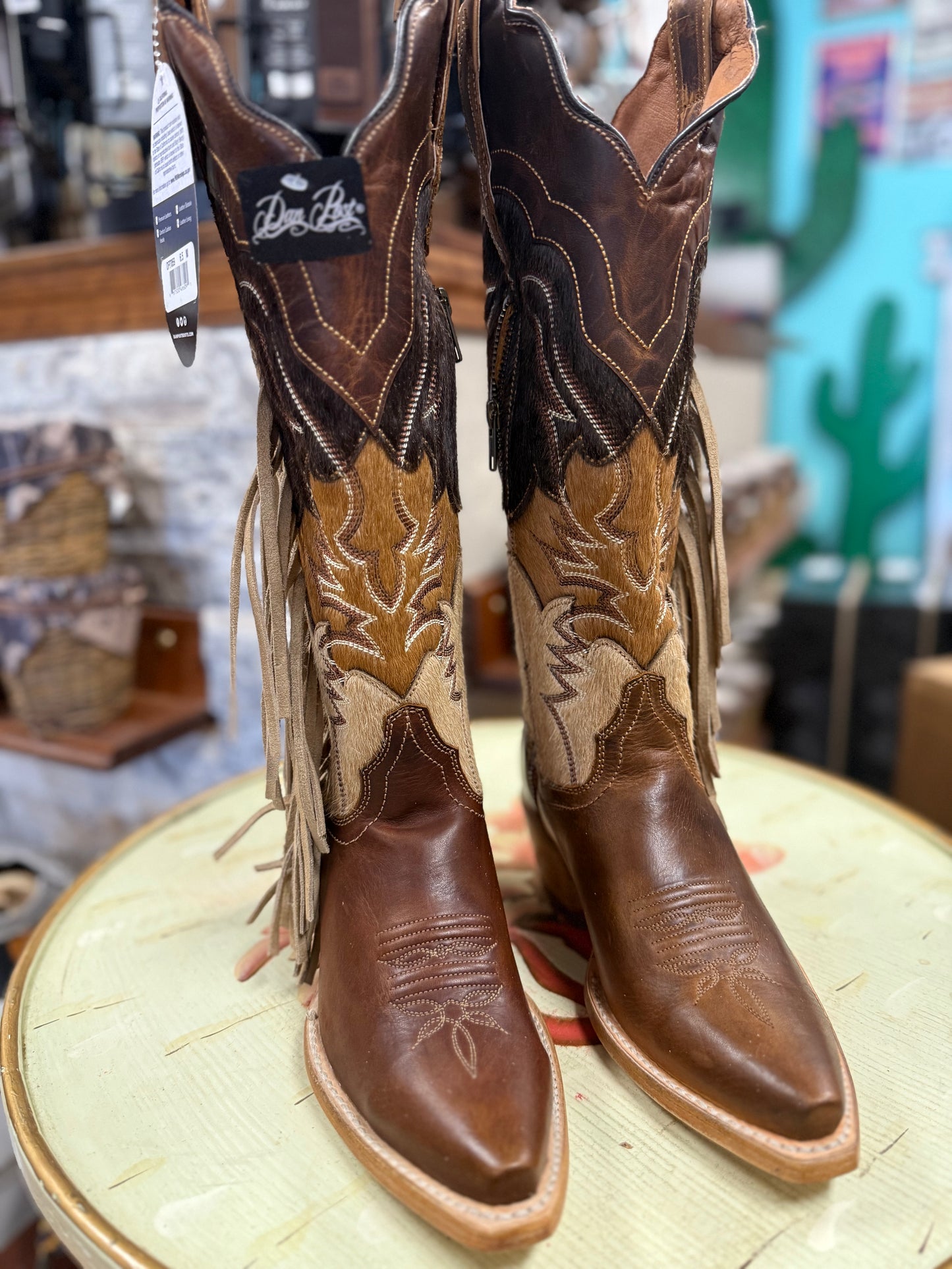 Pair of brown cowboy boots with fringe on a round surface, with a blurred store background.