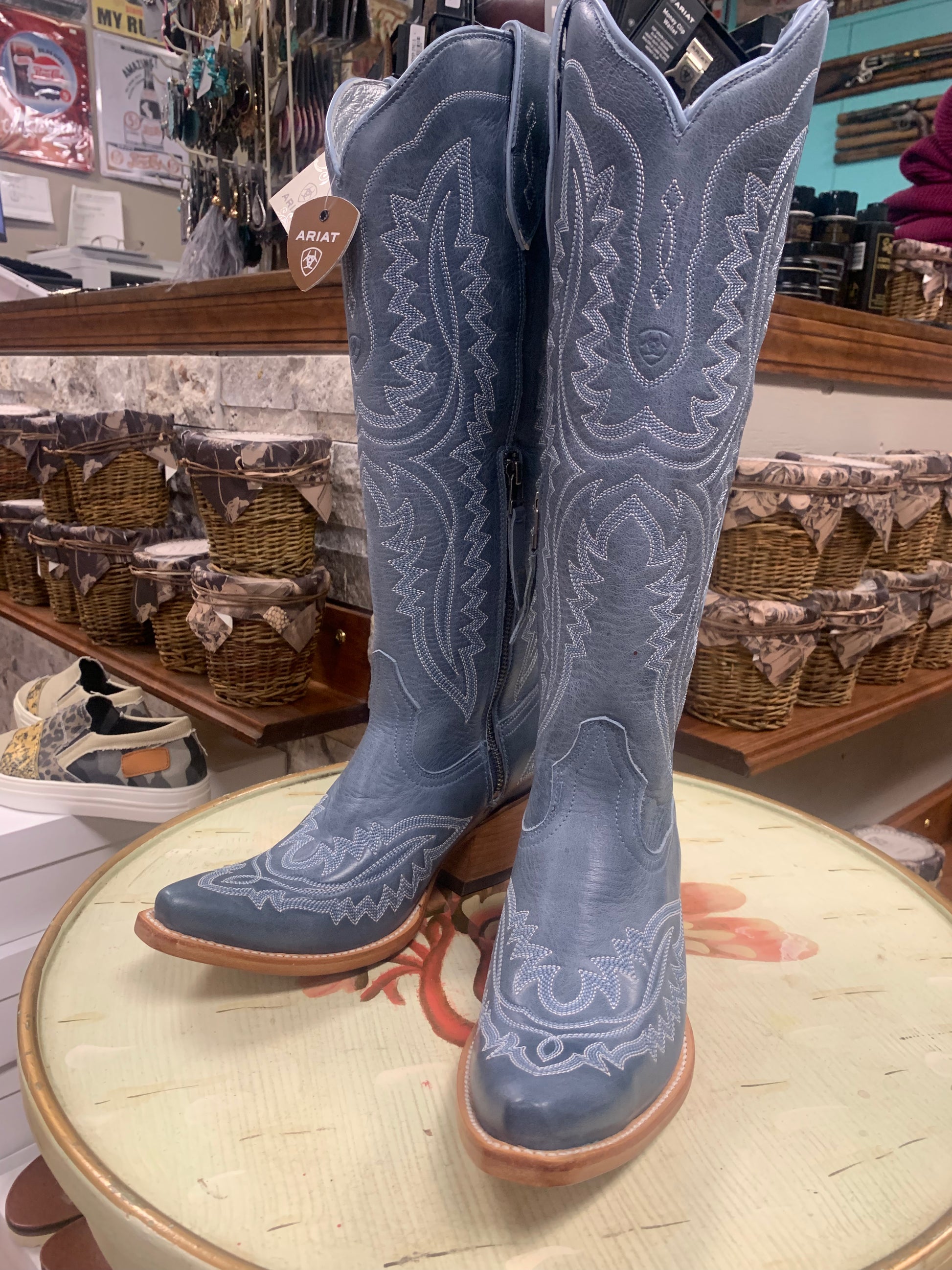 A pair of blue denim western boots with detailed stitching, displayed on a table with a country store background.