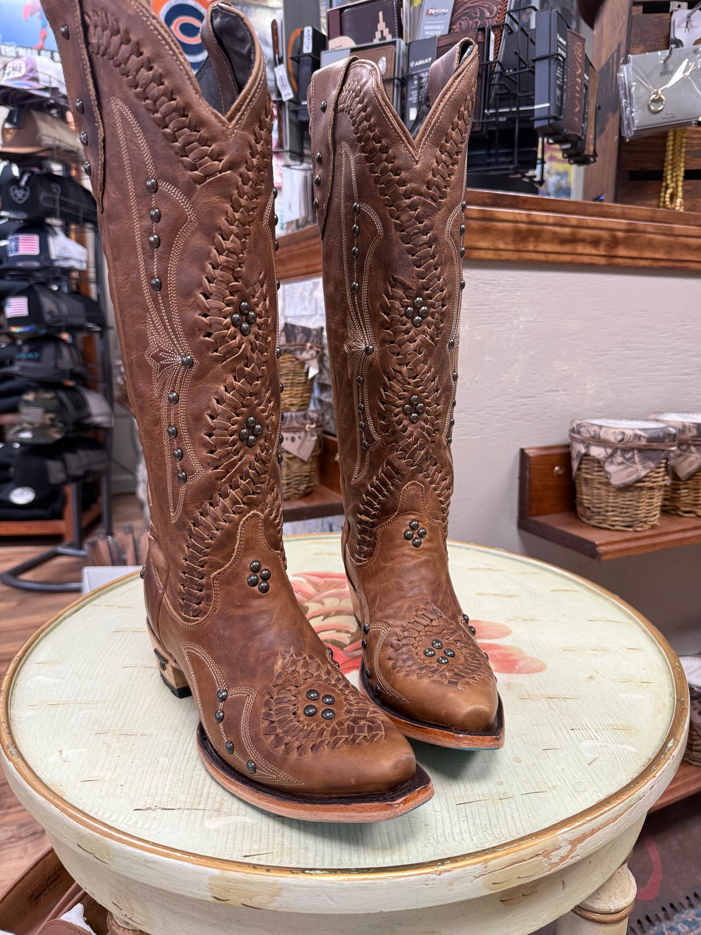 Pair of brown cowboy boots with intricate designs on a round table in a store setting.