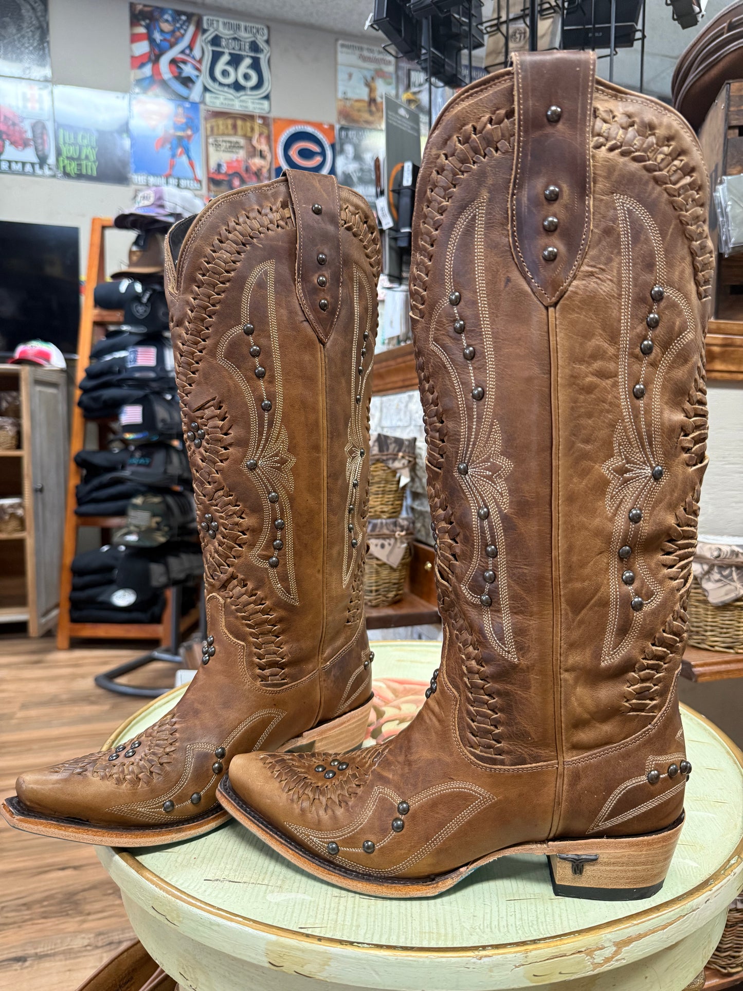 Pair of brown cowboy boots with intricate designs on a round surface, with a store interior in the background.