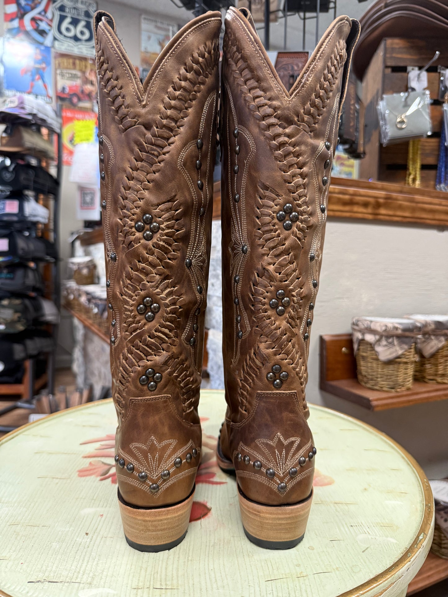 Pair of brown cowboy boots with intricate designs on a round surface in a store setting.