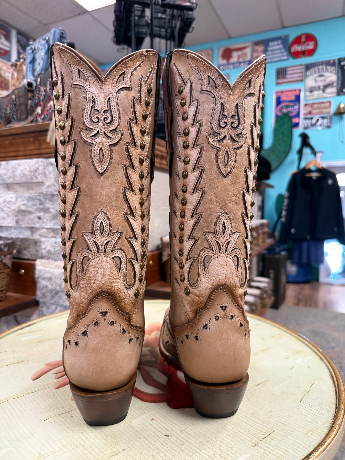 Pair of tan cowboy boots with intricate designs on a wooden surface, with a store interior in the background.