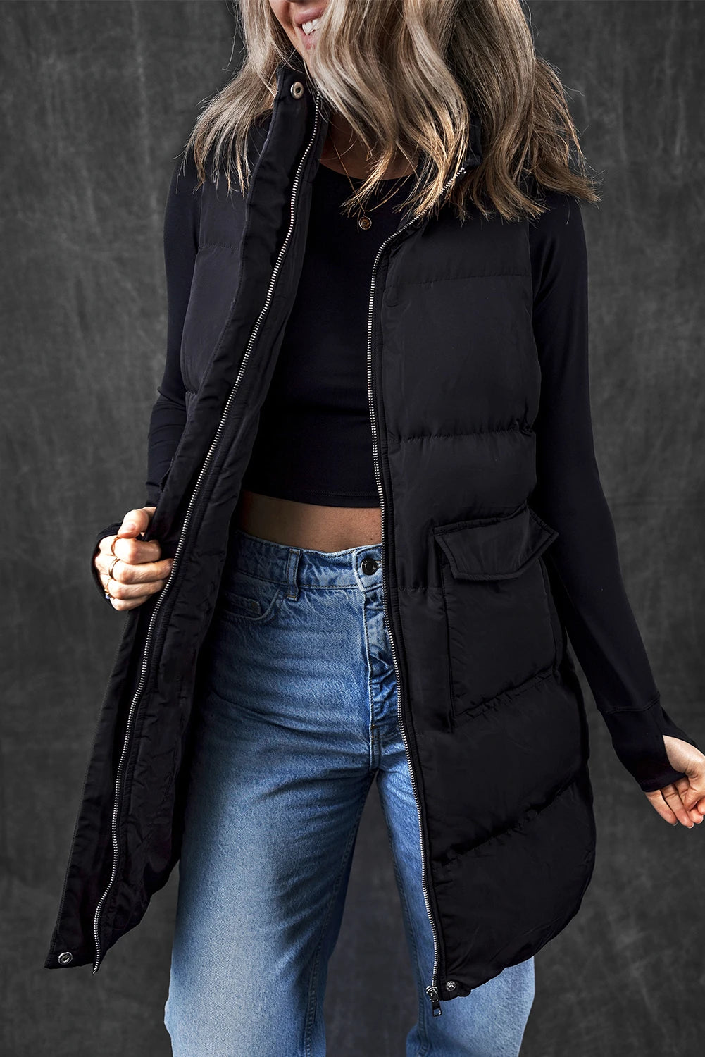 Person wearing a black puffer vest over a black top and blue jeans against a dark background
