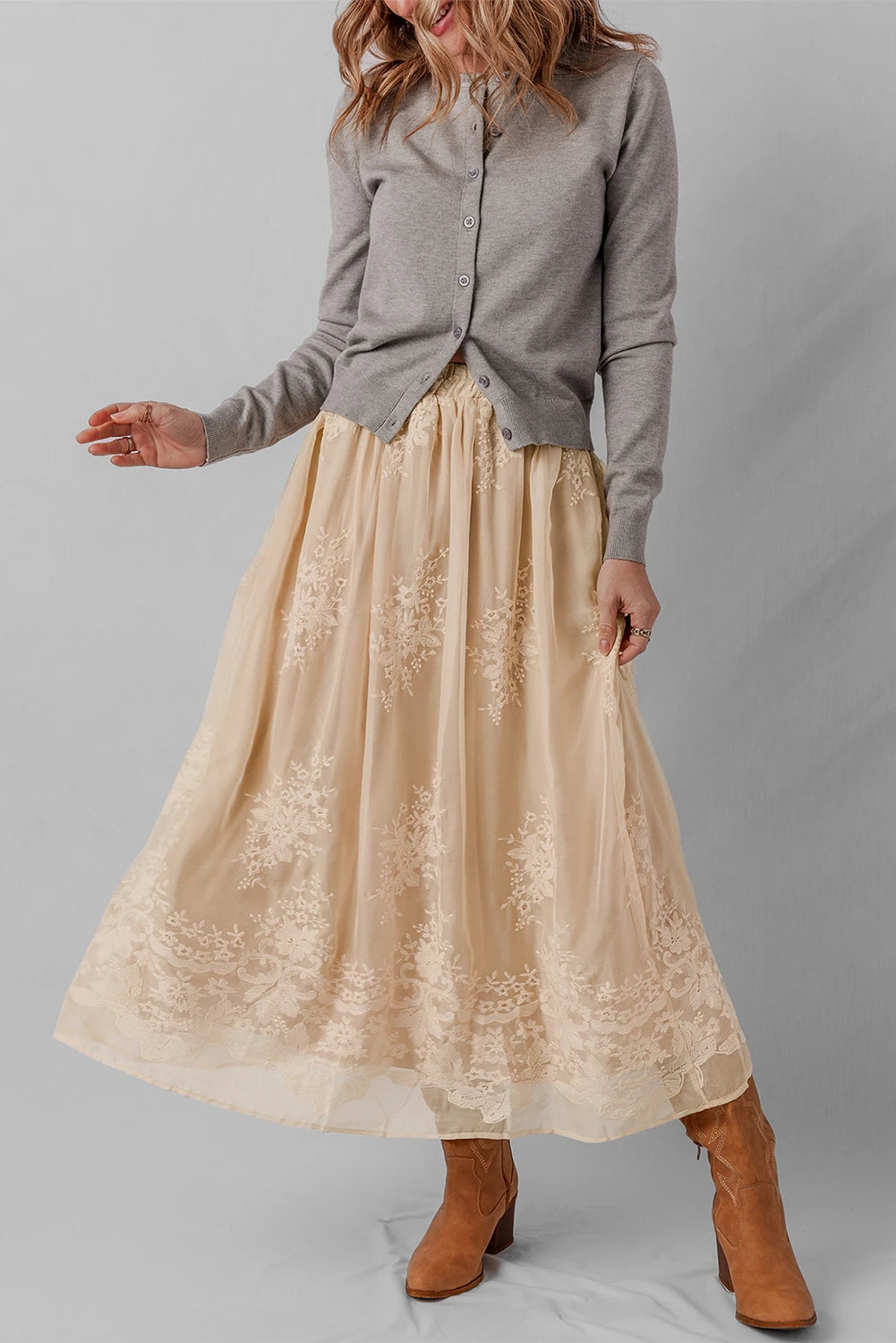 Woman wearing a gray shirt and beige lace skirt with brown boots on a plain background