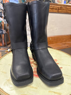 A pair of black leather mid-calf boots with square toes and a wide strap detail around the ankle, displayed on a round wooden table in a store setting.