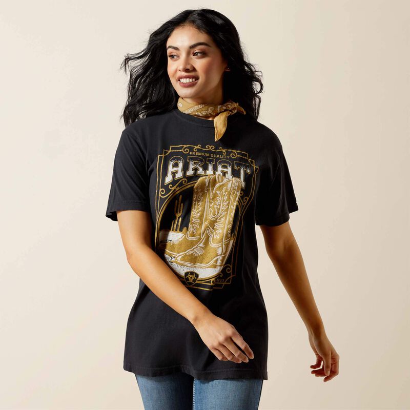 Woman wearing a black t-shirt with gold graphics and 'Ariat' branding on a beige background