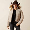 Woman wearing a beige puffer jacket, black top, dark jeans, and a brown cowboy hat on a beige background