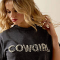 Woman wearing a black t-shirt with 'COWGIRL' in pearls, posing with one hand in her hair.