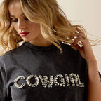 Woman wearing a black t-shirt with 'COWGIRL' in pearls, posing with one hand in her hair.
