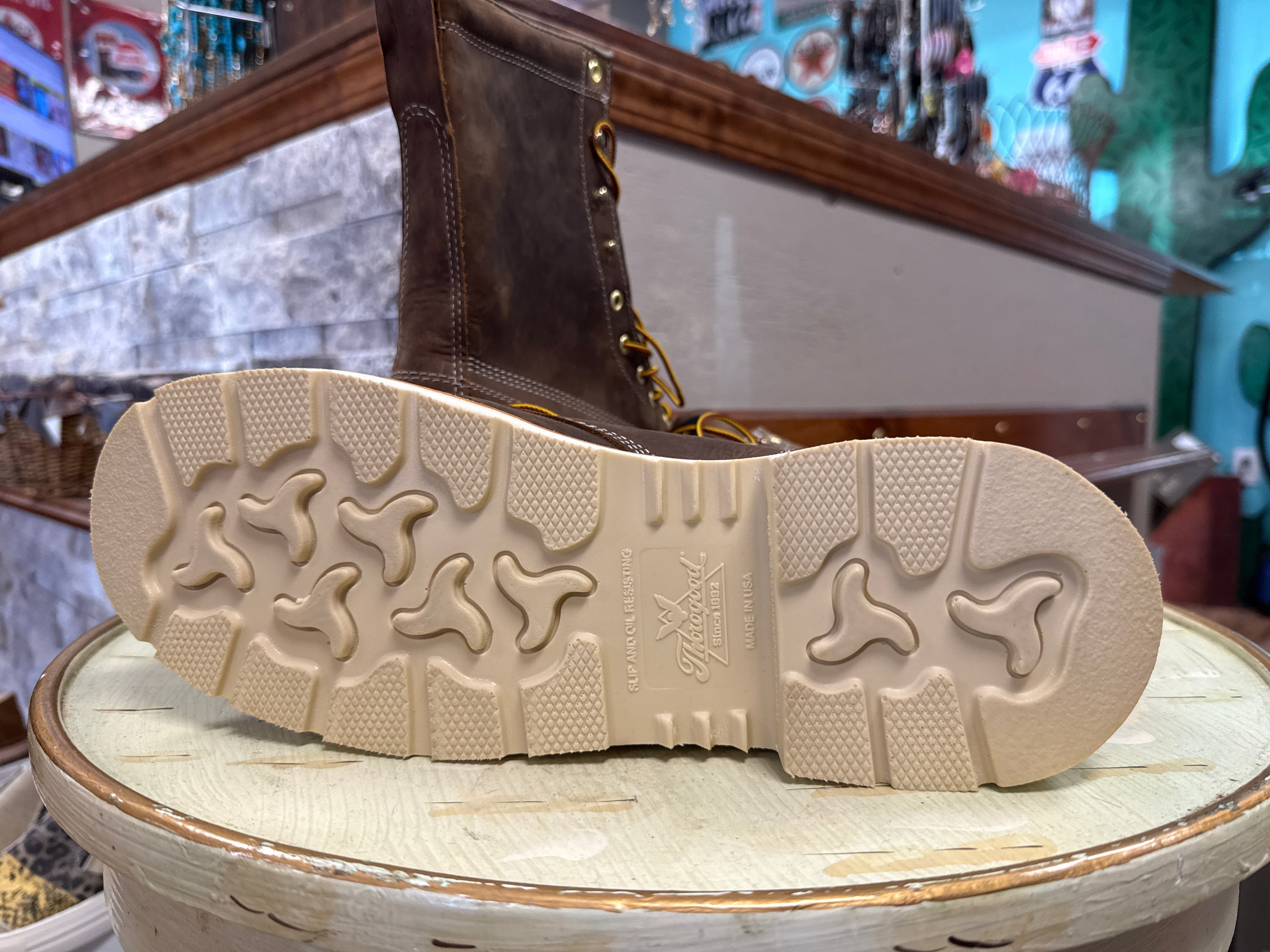 A close-up of a brown leather lace-up boot shown from the bottom, highlighting its beige treaded sole, displayed on a rustic wooden stand in a store setting.