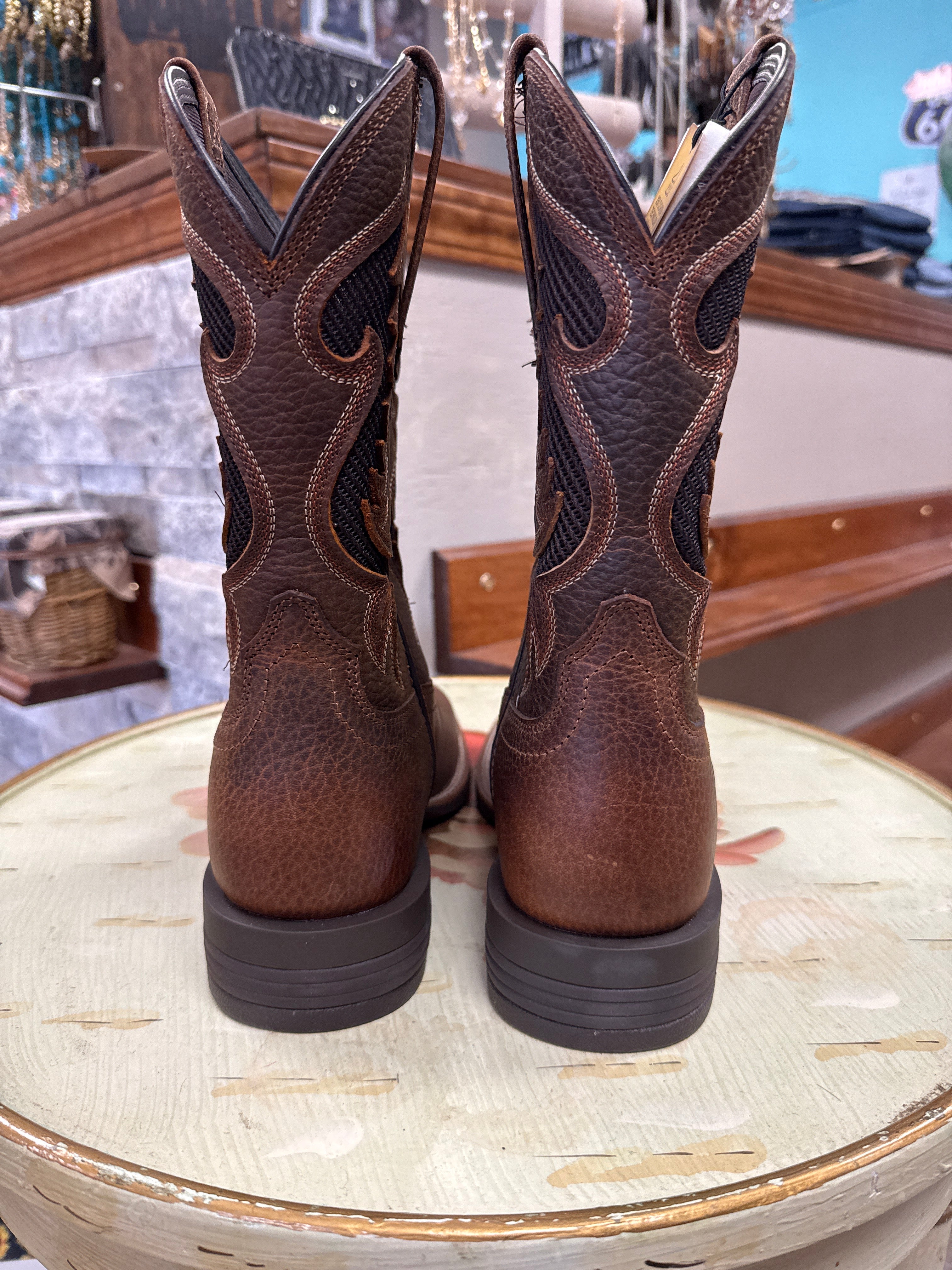 A pair of dark brown leather cowboy boots with decorative cutout stitching, shown from the back on a rustic wooden table inside a store.