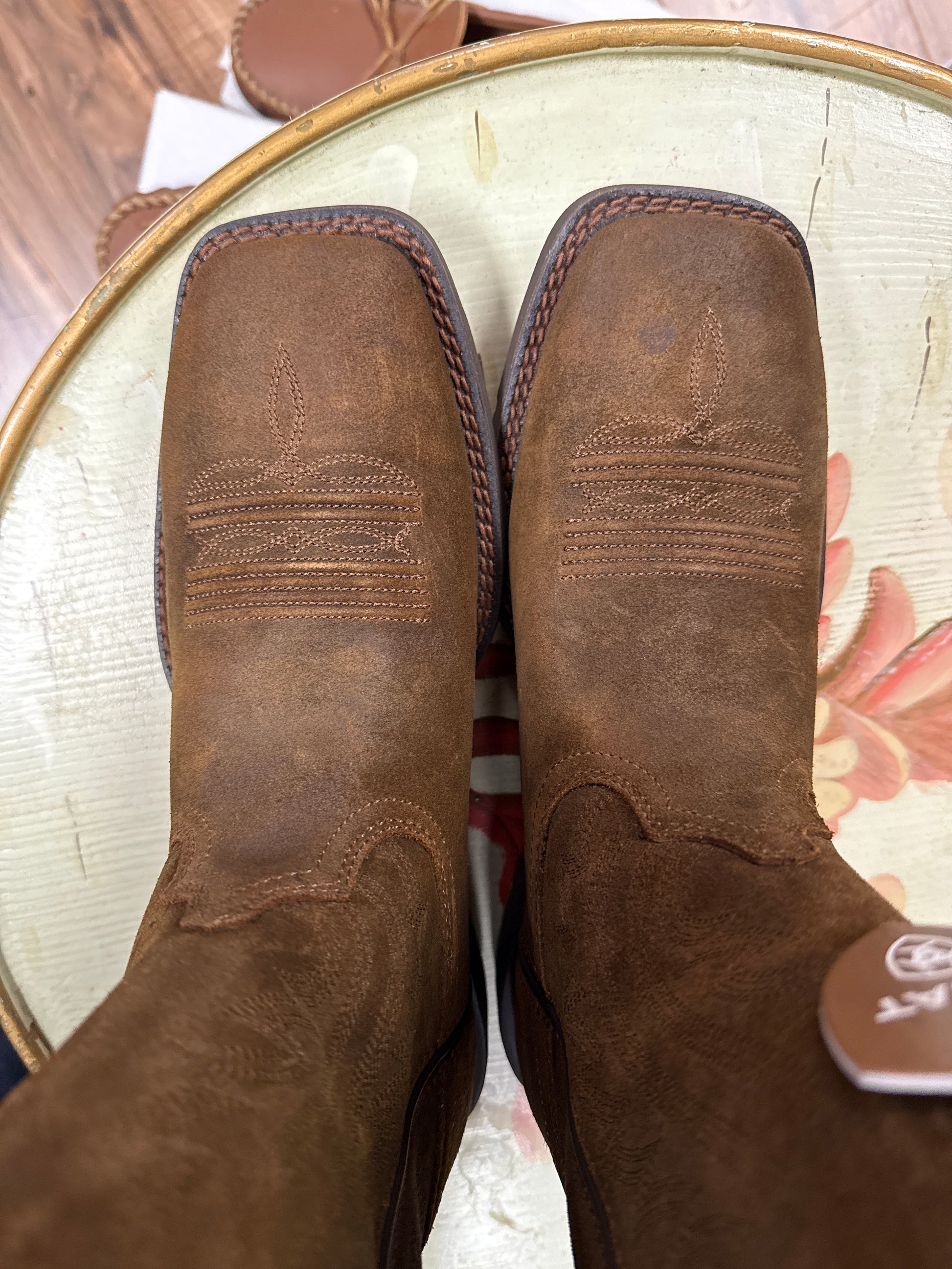 Pair of brown leather boots on a round surface with a floral patterned rug in the background.