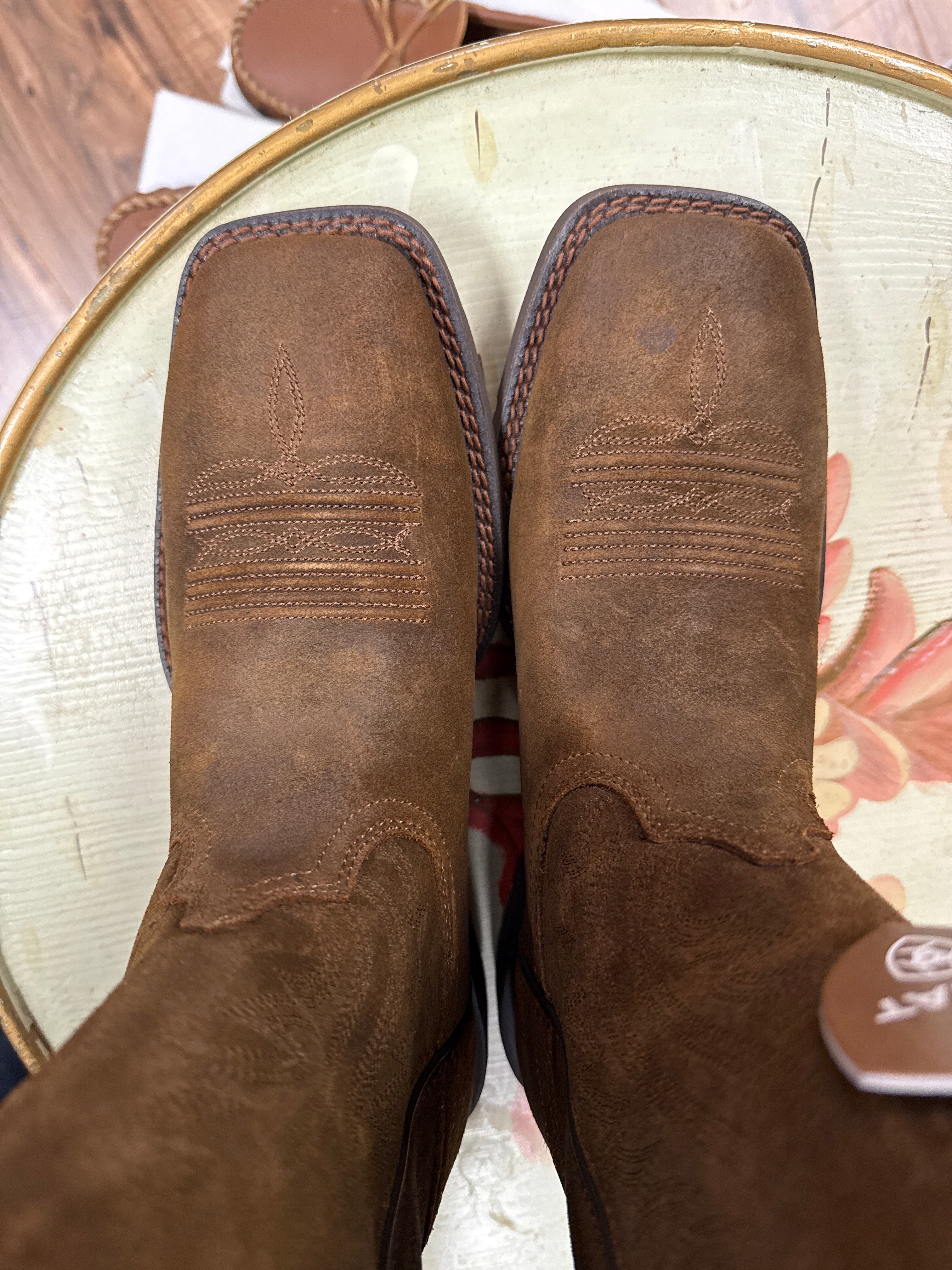 Pair of brown leather boots on a round surface with a floral patterned rug in the background.