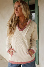 Woman wearing a cream and pink sweater with a textured pattern, standing against a rustic wall.