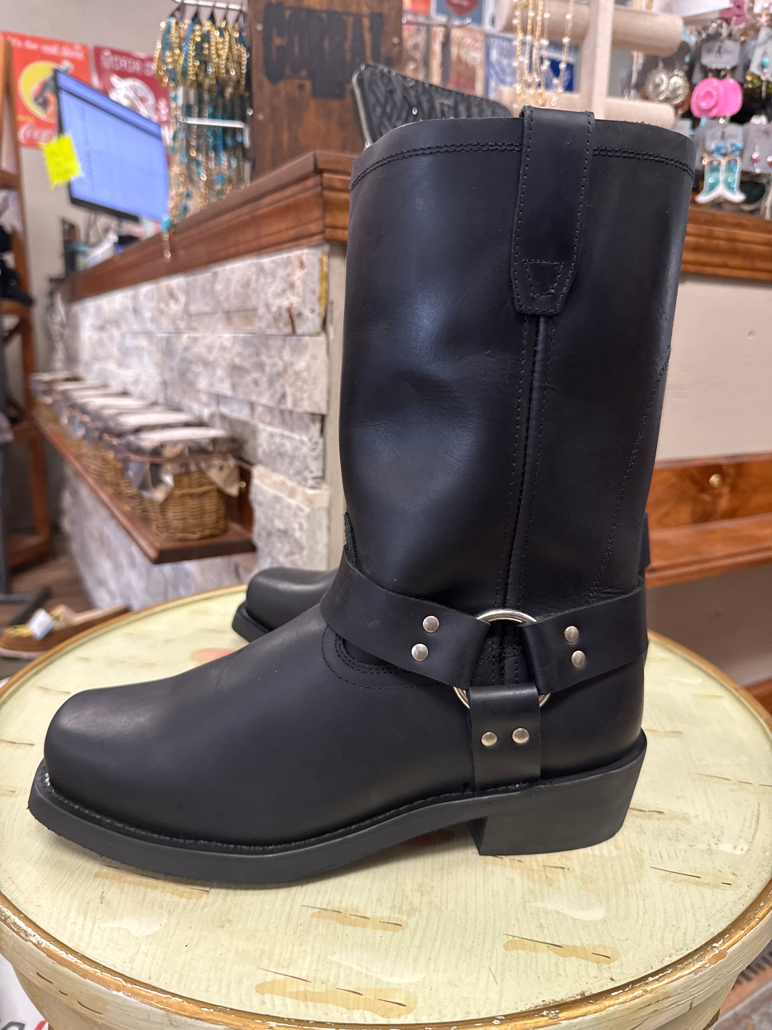 A pair of black leather mid-calf boots with a buckle strap detail, displayed on a round wooden table inside a retail store.