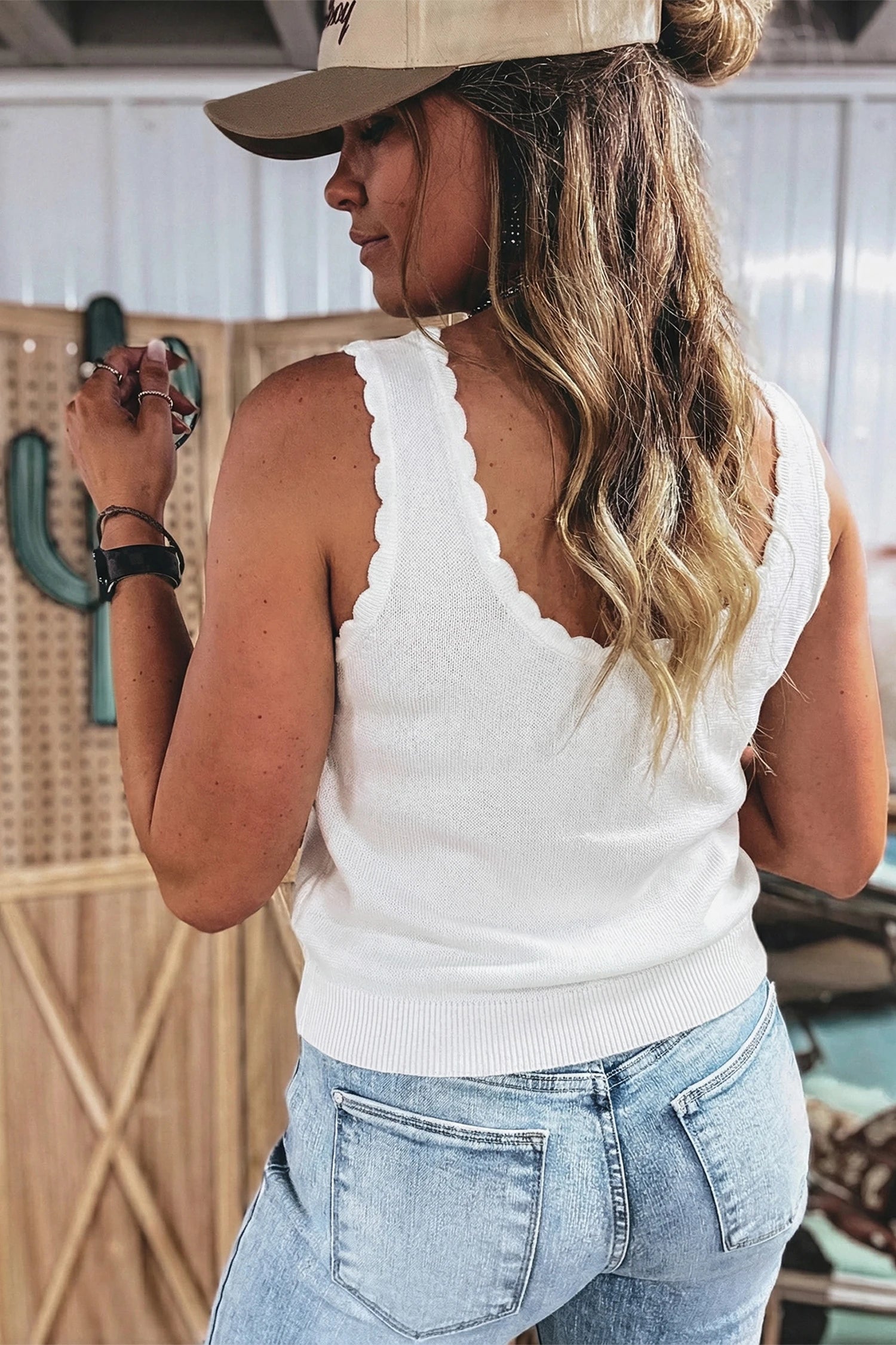 Woman wearing a white sleeveless top and blue jeans with a beige cap indoors.