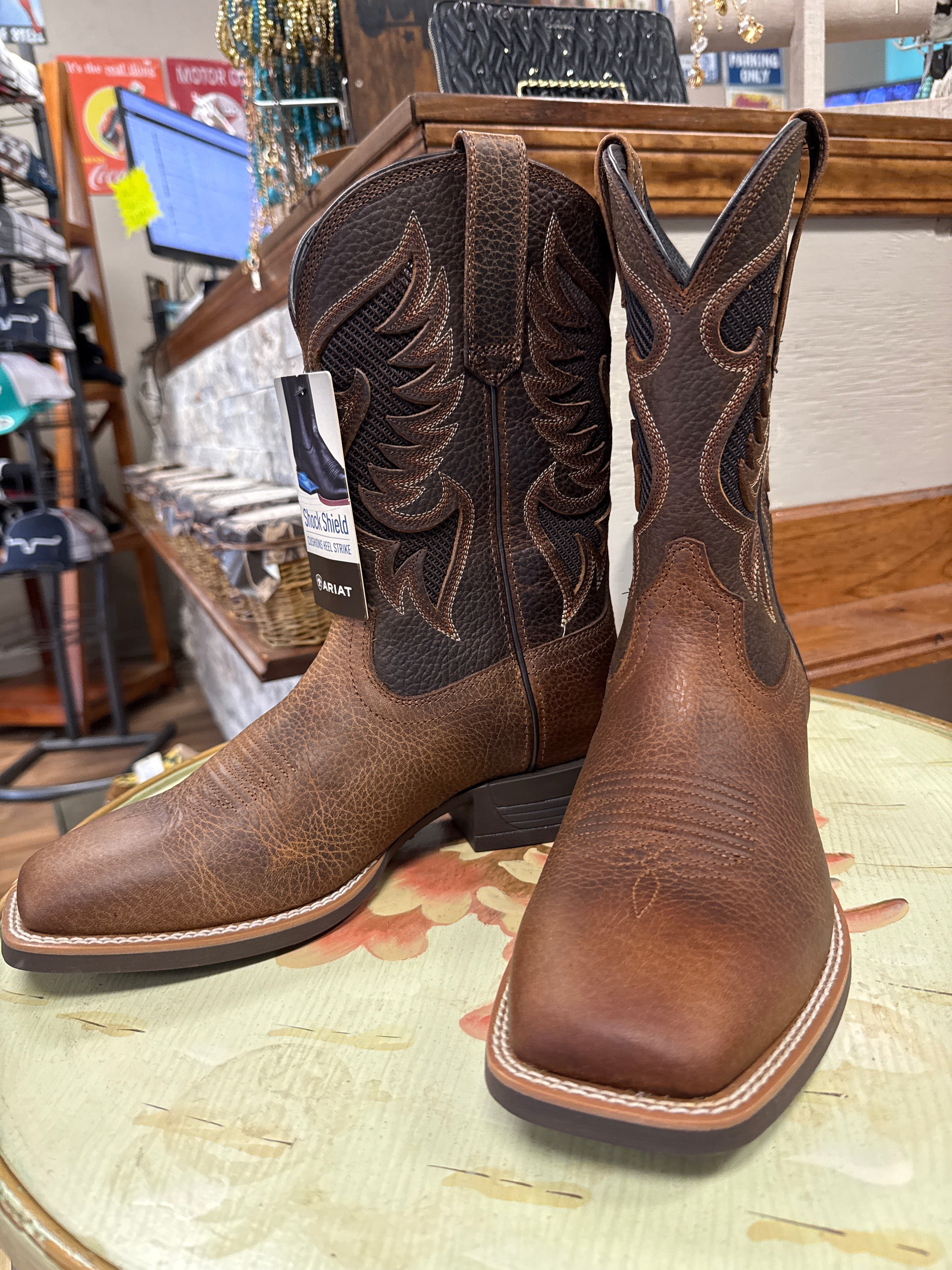 A pair of western cowboy boots in brown leather, displayed on a round table inside a retail store. The boots feature dark brown embroidered stitching on the shafts and a lighter brown toe and vamp.