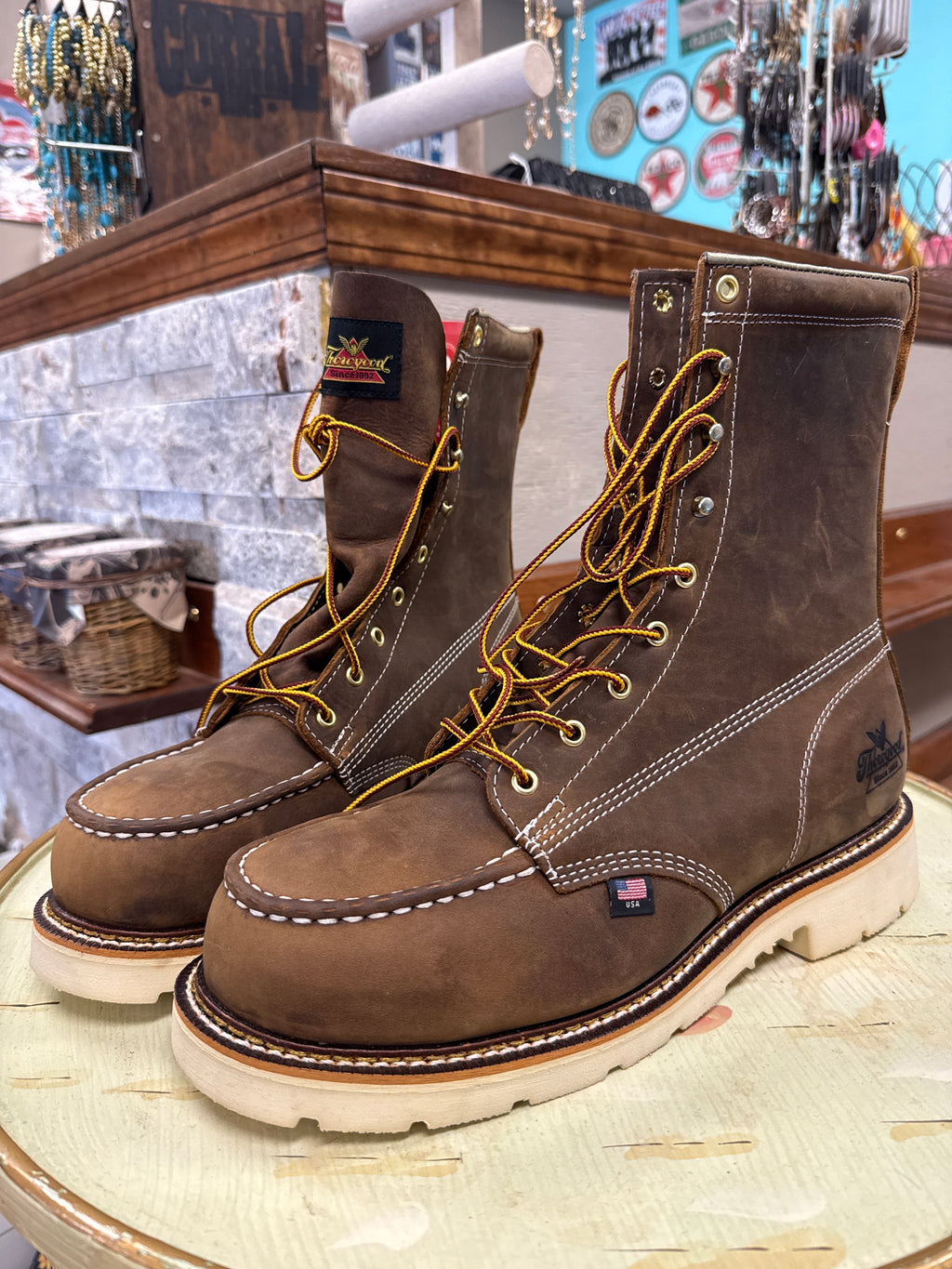 A pair of brown leather lace-up work boots with white contrast stitching and yellow laces, displayed on a light-colored wooden table inside a store.