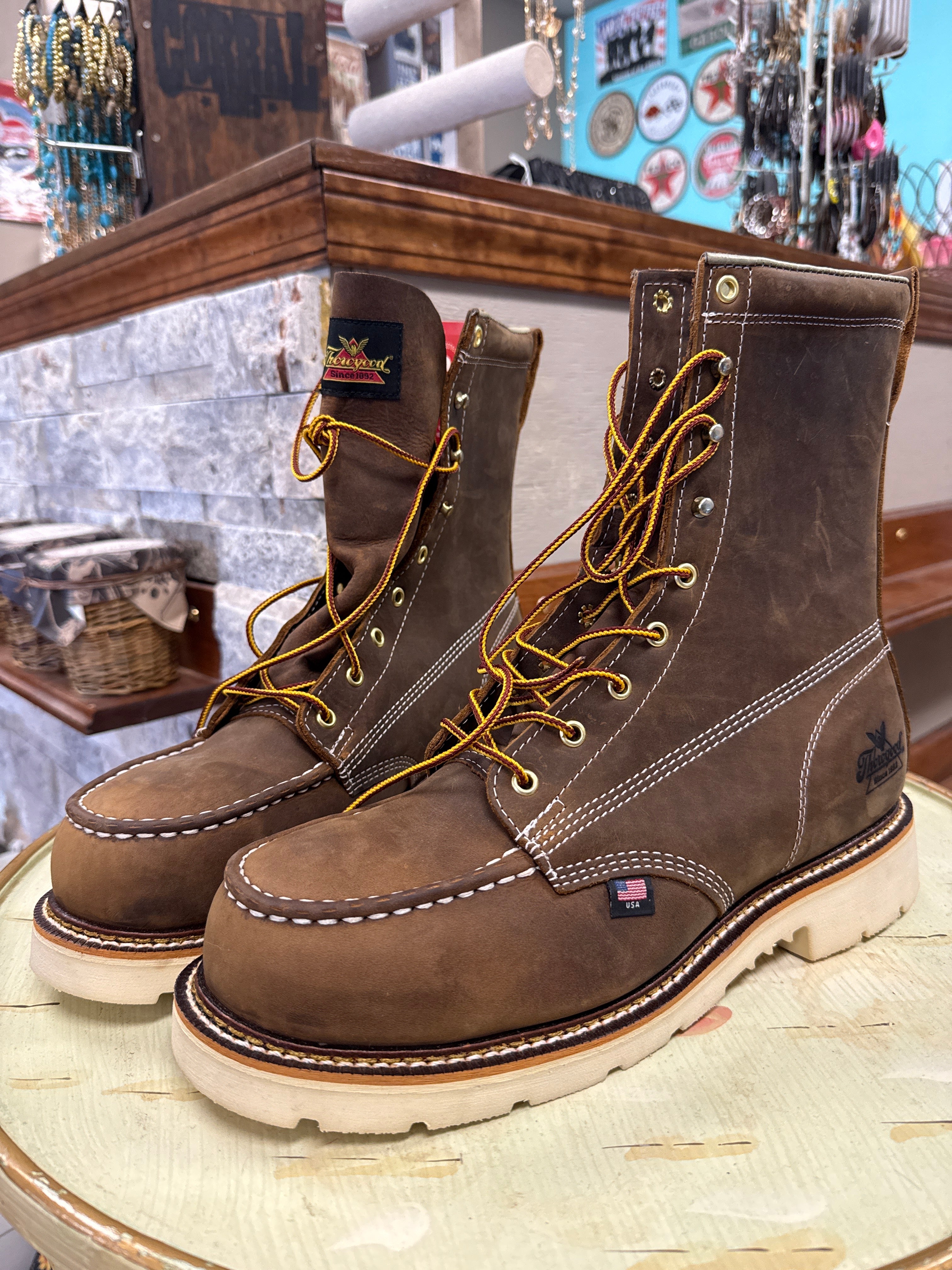 A pair of brown leather lace-up work boots with white contrast stitching and yellow laces, displayed on a light-colored wooden table inside a store.