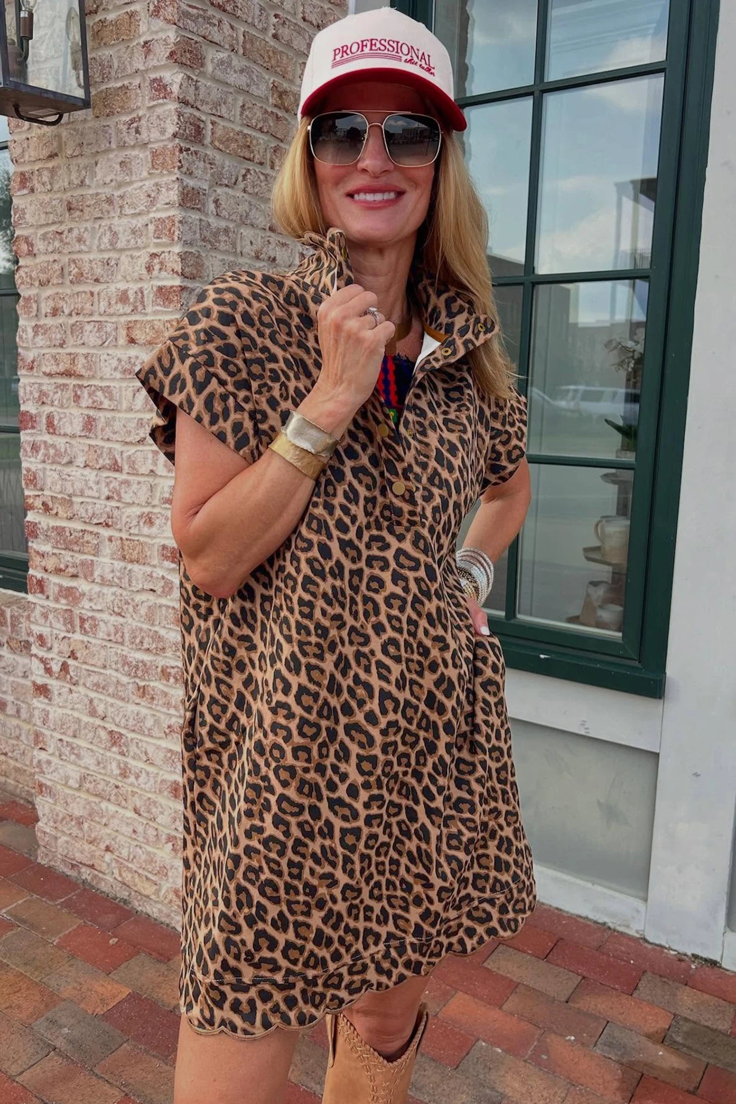 Woman wearing a leopard print dress and red cap with sunglasses, standing against a brick wall.