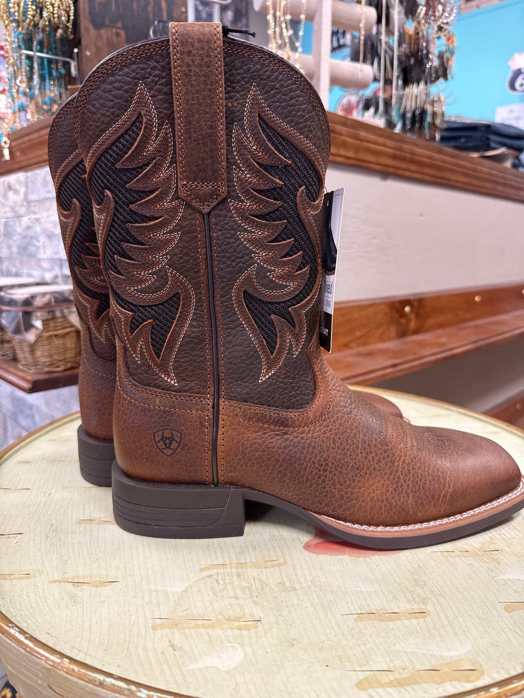 A pair of Ariat brown leather cowboy boots with dark brown western embroidery on the shaft, displayed on a light-colored wooden stool inside a store.