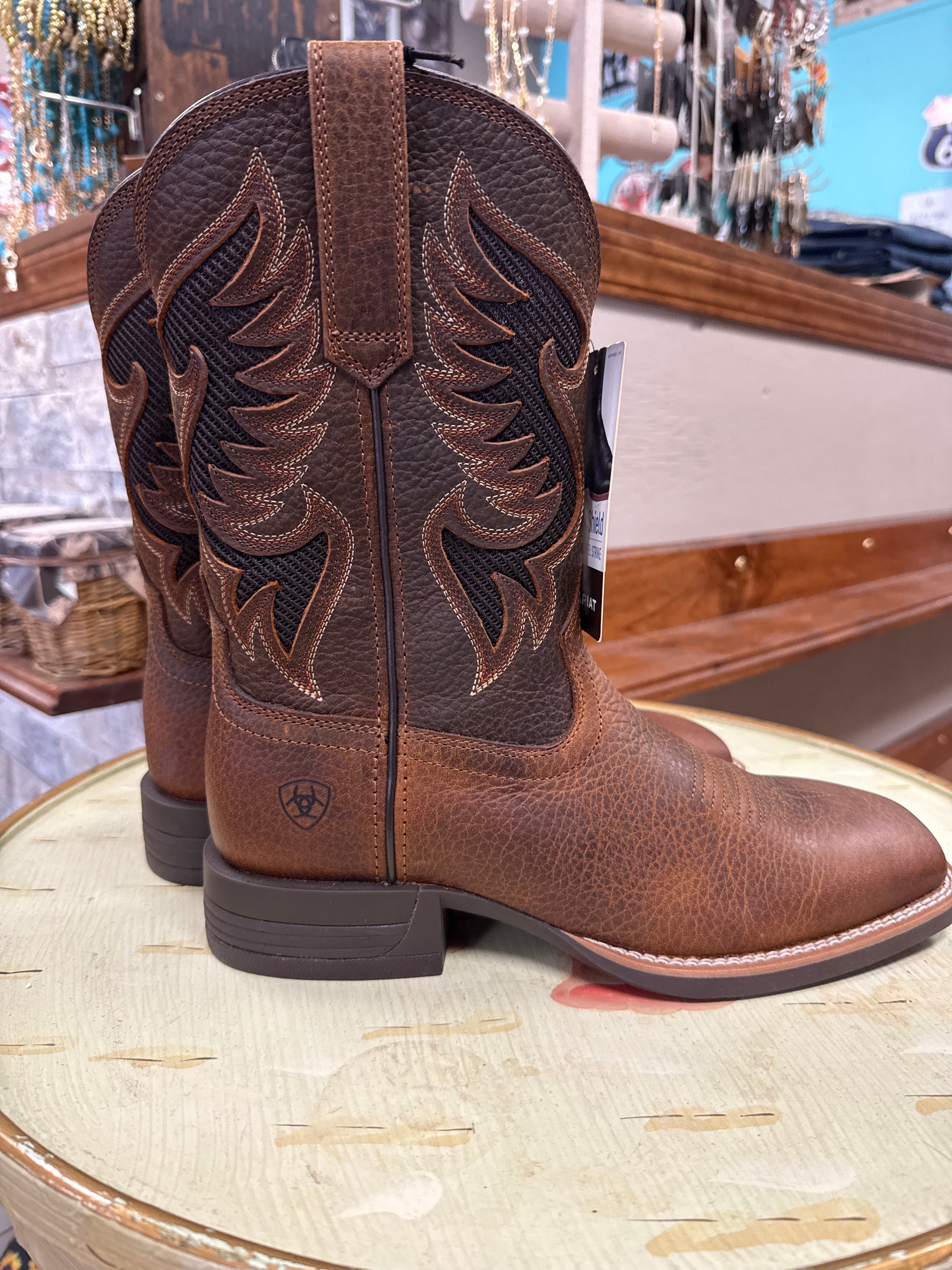 A pair of Ariat brown leather cowboy boots with dark brown western embroidery on the shaft, displayed on a light-colored wooden stool inside a store.