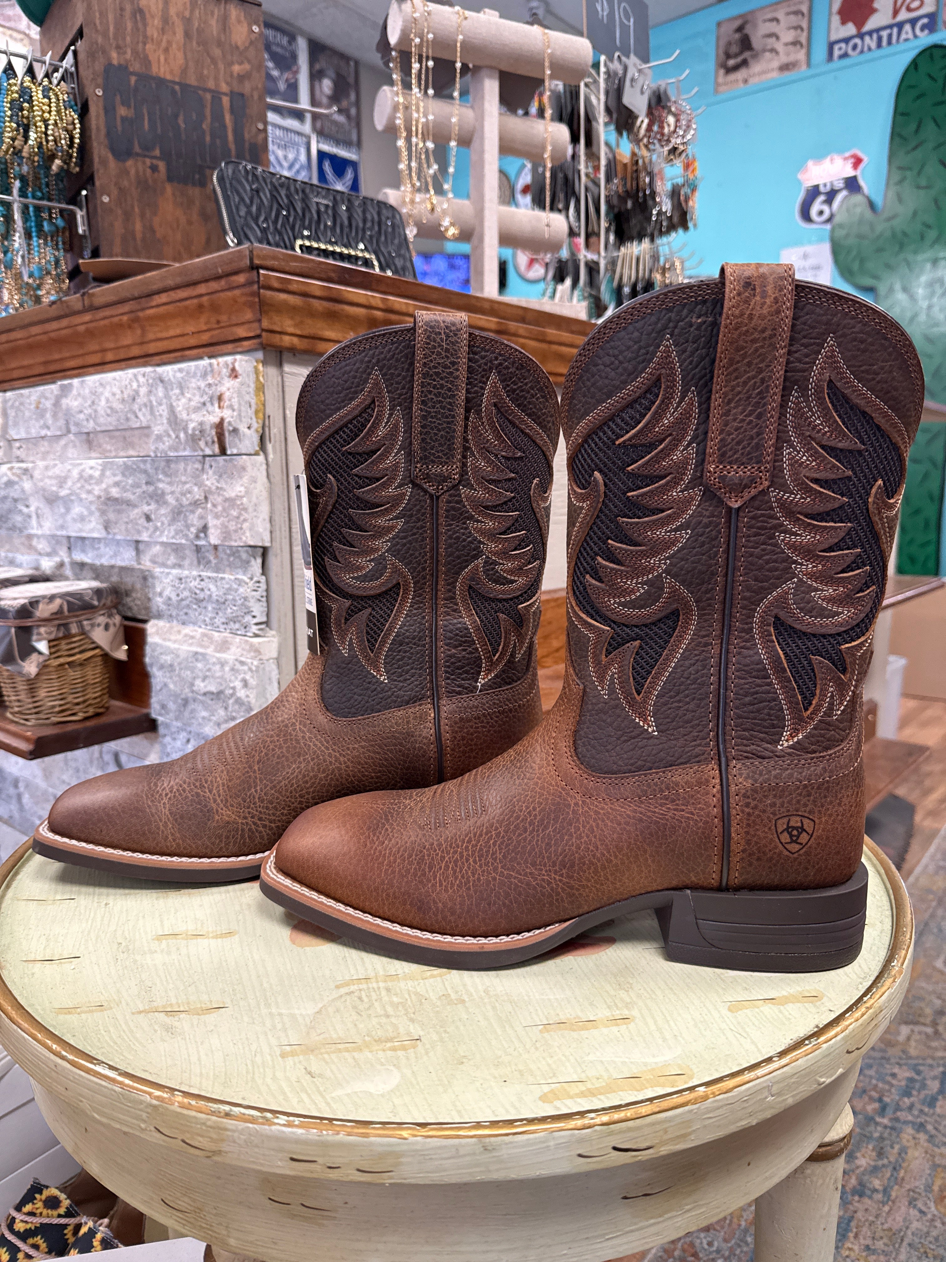 A pair of brown leather western boots with dark brown embroidered shafts, displayed on a distressed white wooden stool inside a retail store.