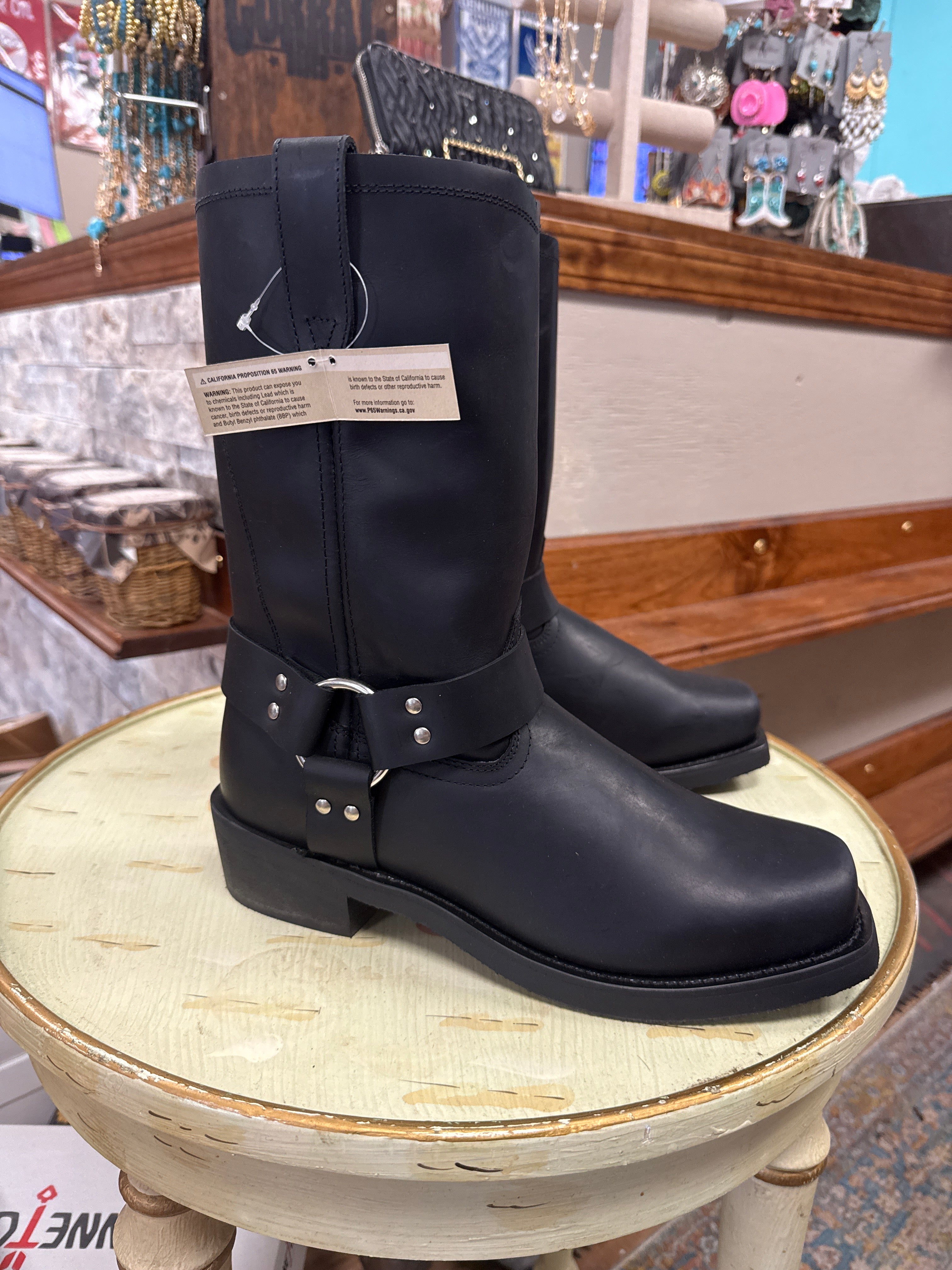 A pair of black leather mid-calf boots with buckle straps, displayed on a distressed wooden stool inside a retail store.