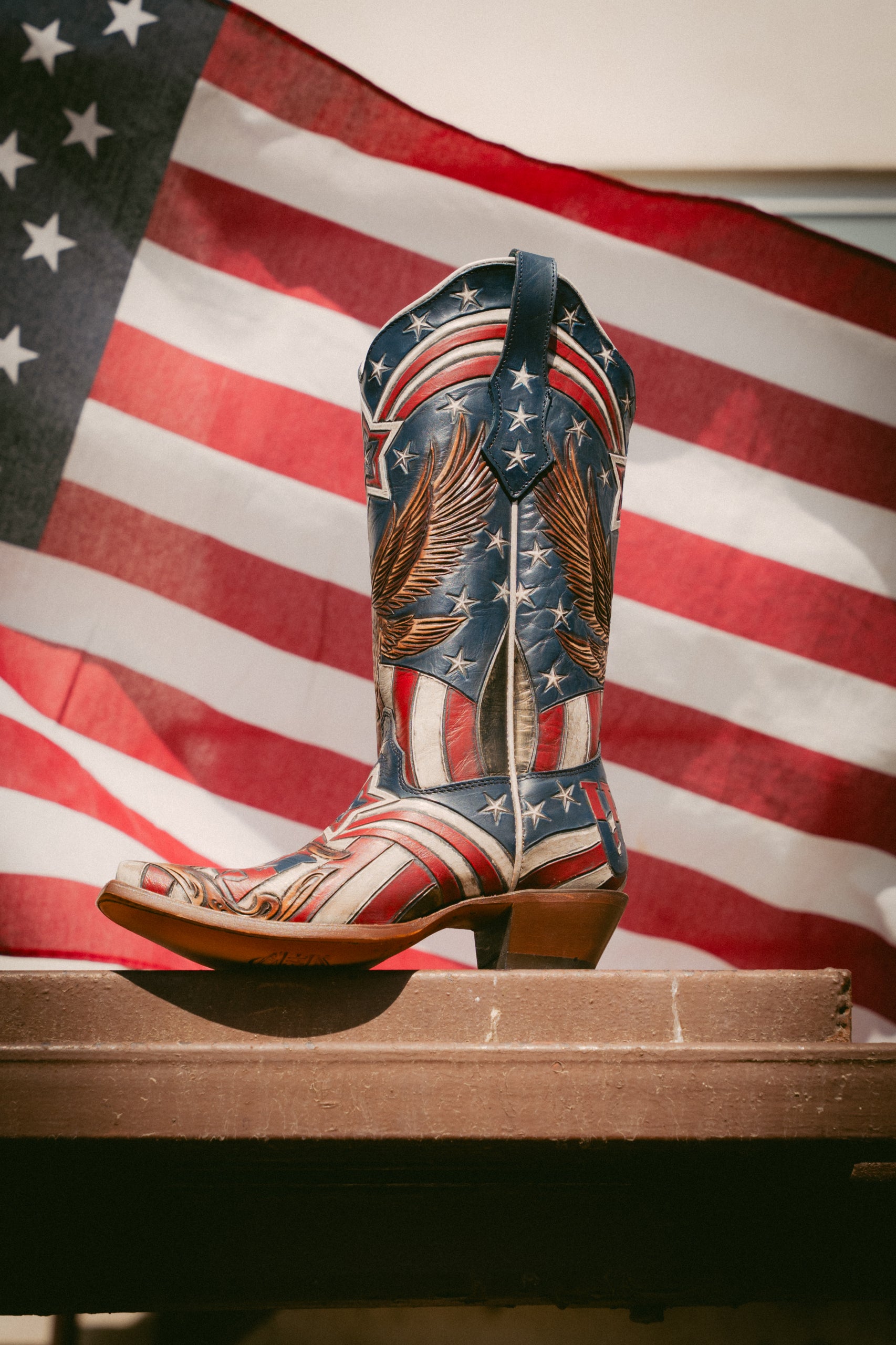 Decorative cowboy boot with American flag design on a wooden surface in front of an American flag.