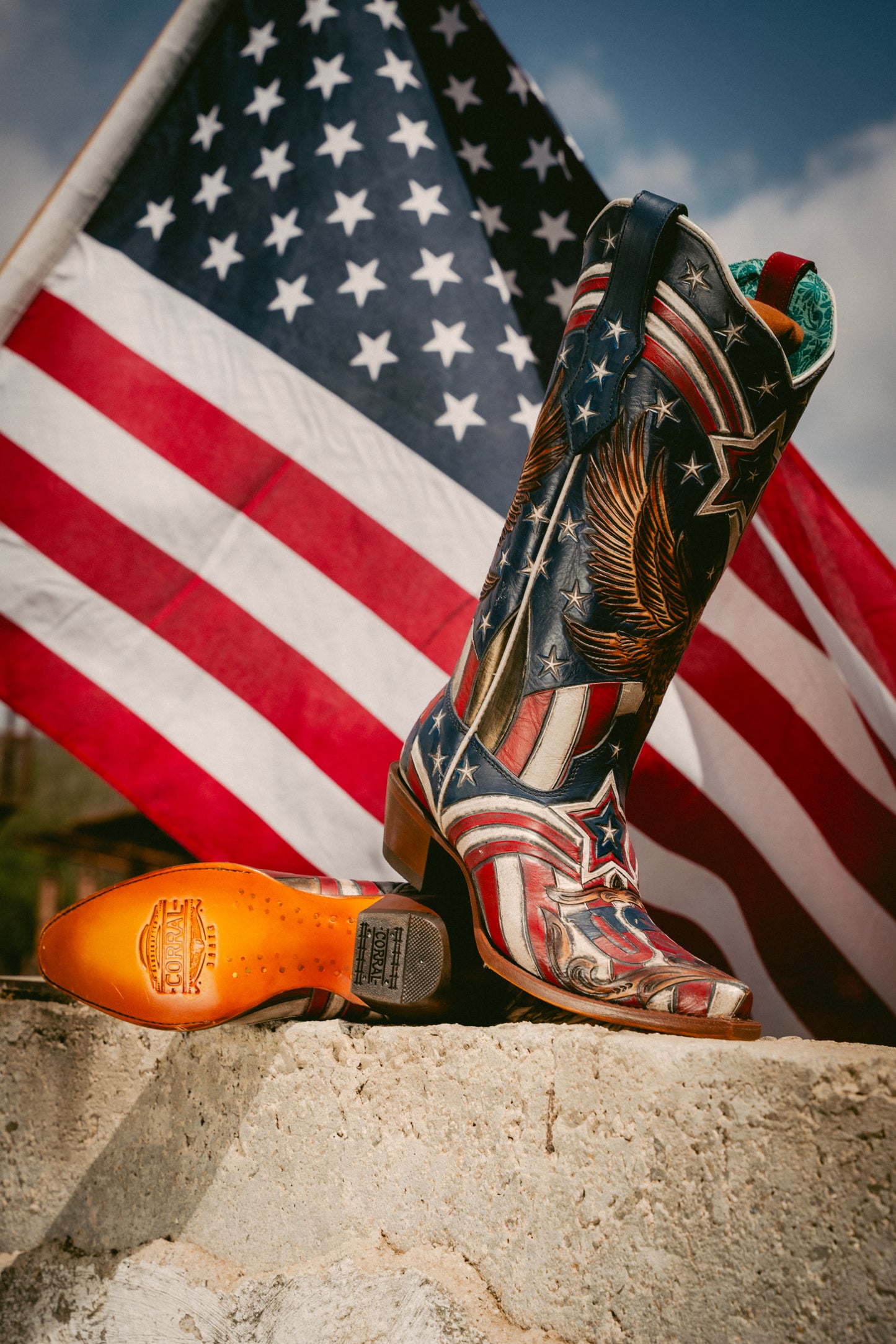 Colorful cowboy boot with American flag design on a concrete surface with an American flag in the background.