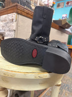 A black leather ankle boot with a buckle strap and textured sole, resting on a distressed cream-colored stool inside a shop.