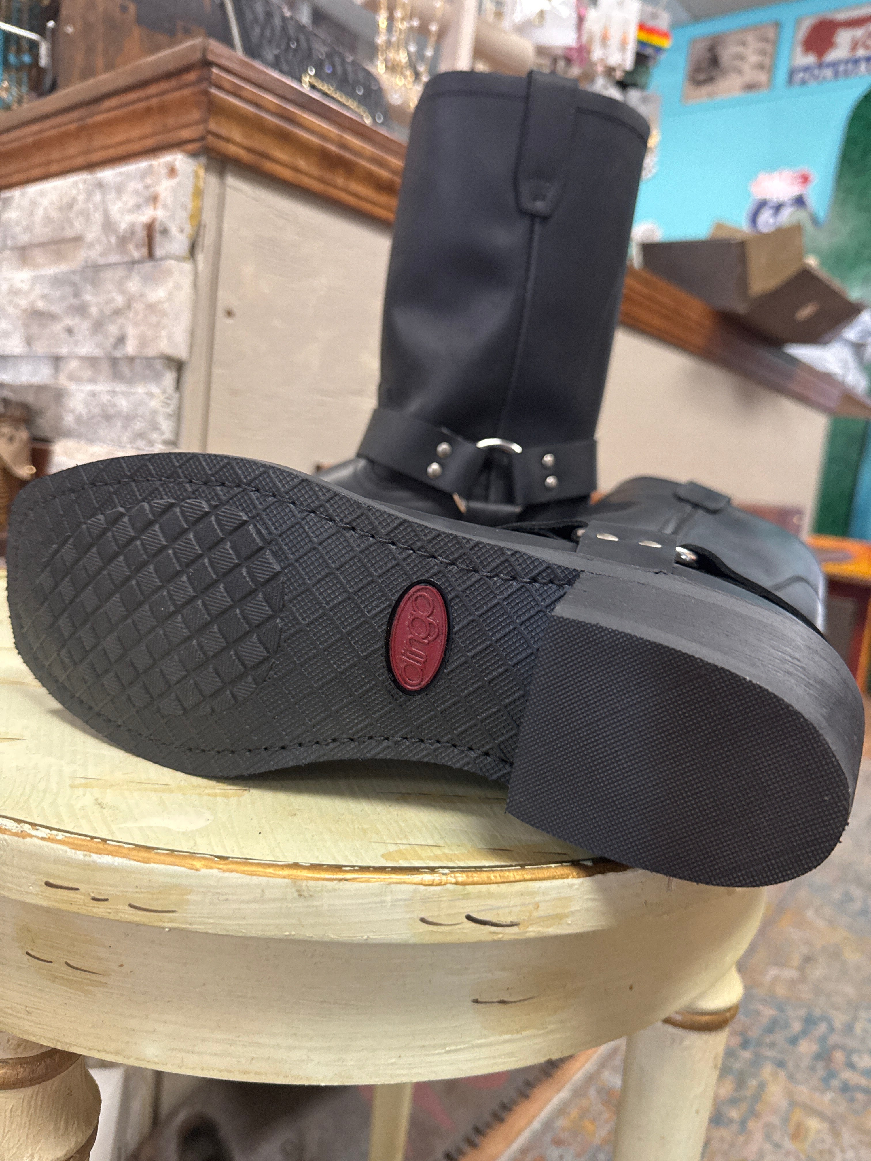 A black leather ankle boot with a buckle strap and textured sole, resting on a distressed cream-colored stool inside a shop.