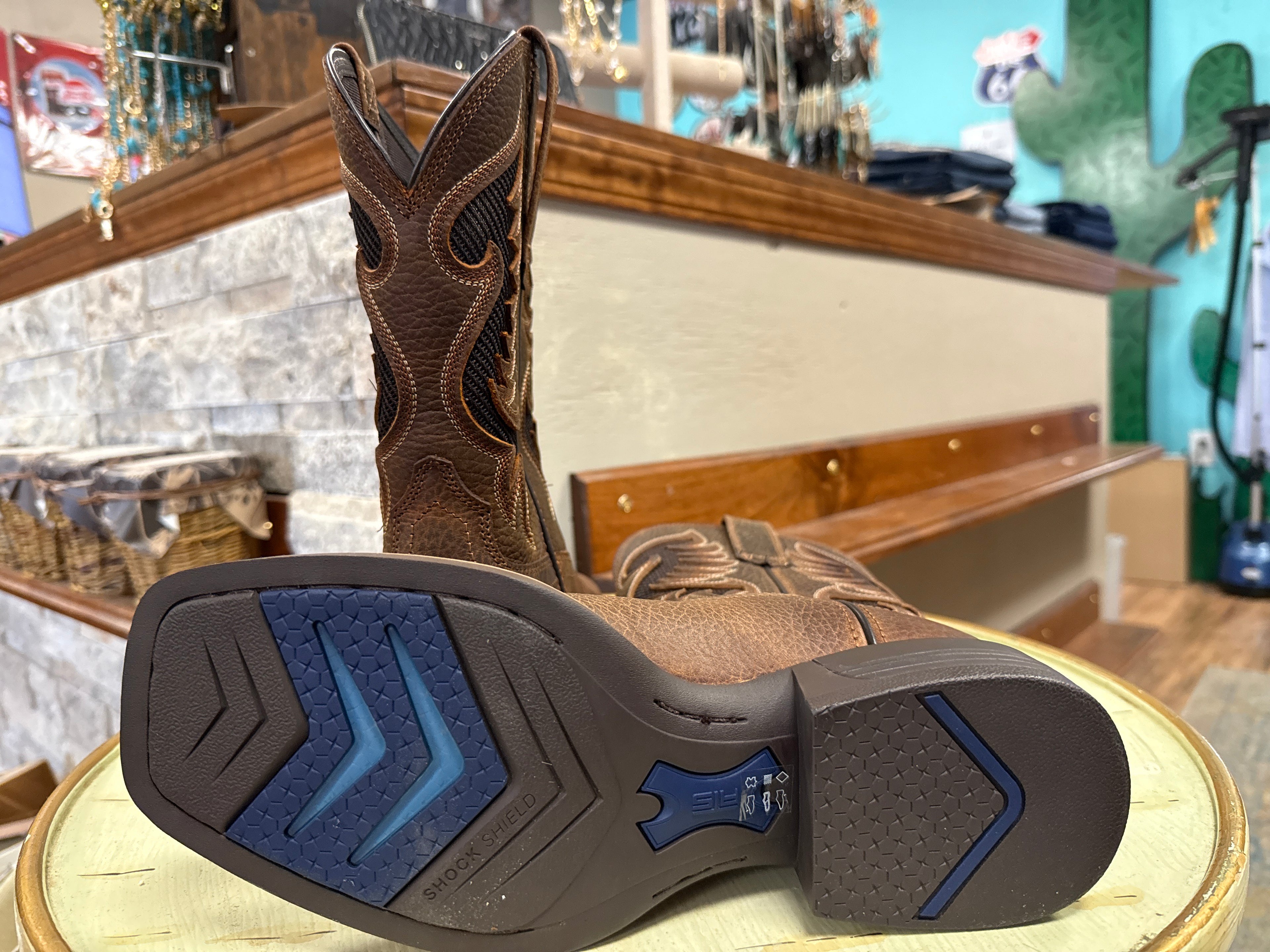 A pair of brown western cowboy boots displayed on a round wooden pedestal inside a retail store. One boot is standing upright to show the shaft, while the other lies flat on its sole to display the tread pattern, which features a dark brown base with blue chevron accents.