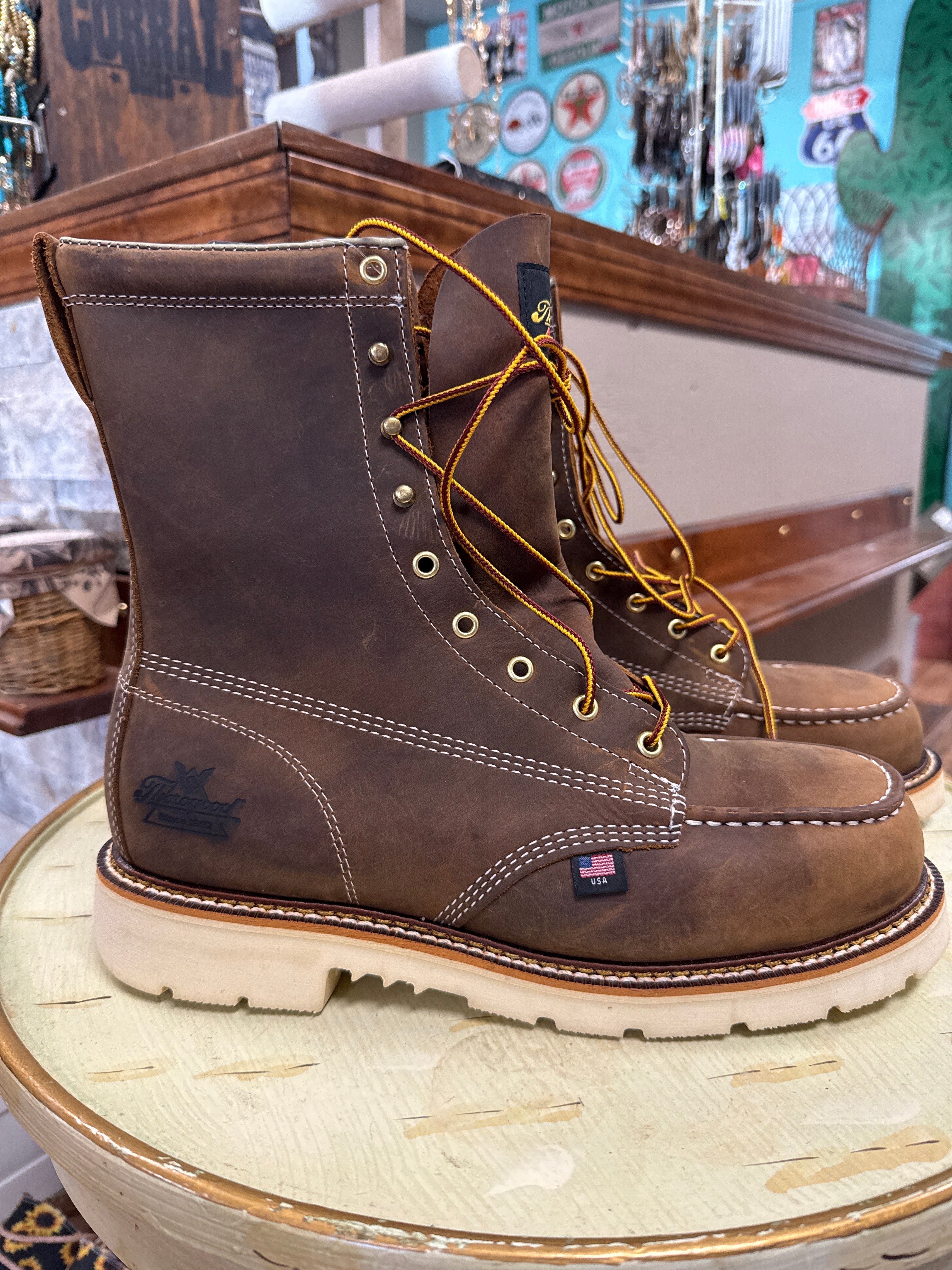 A pair of dark brown leather lace-up work boots with white contrast stitching and tan laces, displayed on a rustic wooden stool inside a shop.