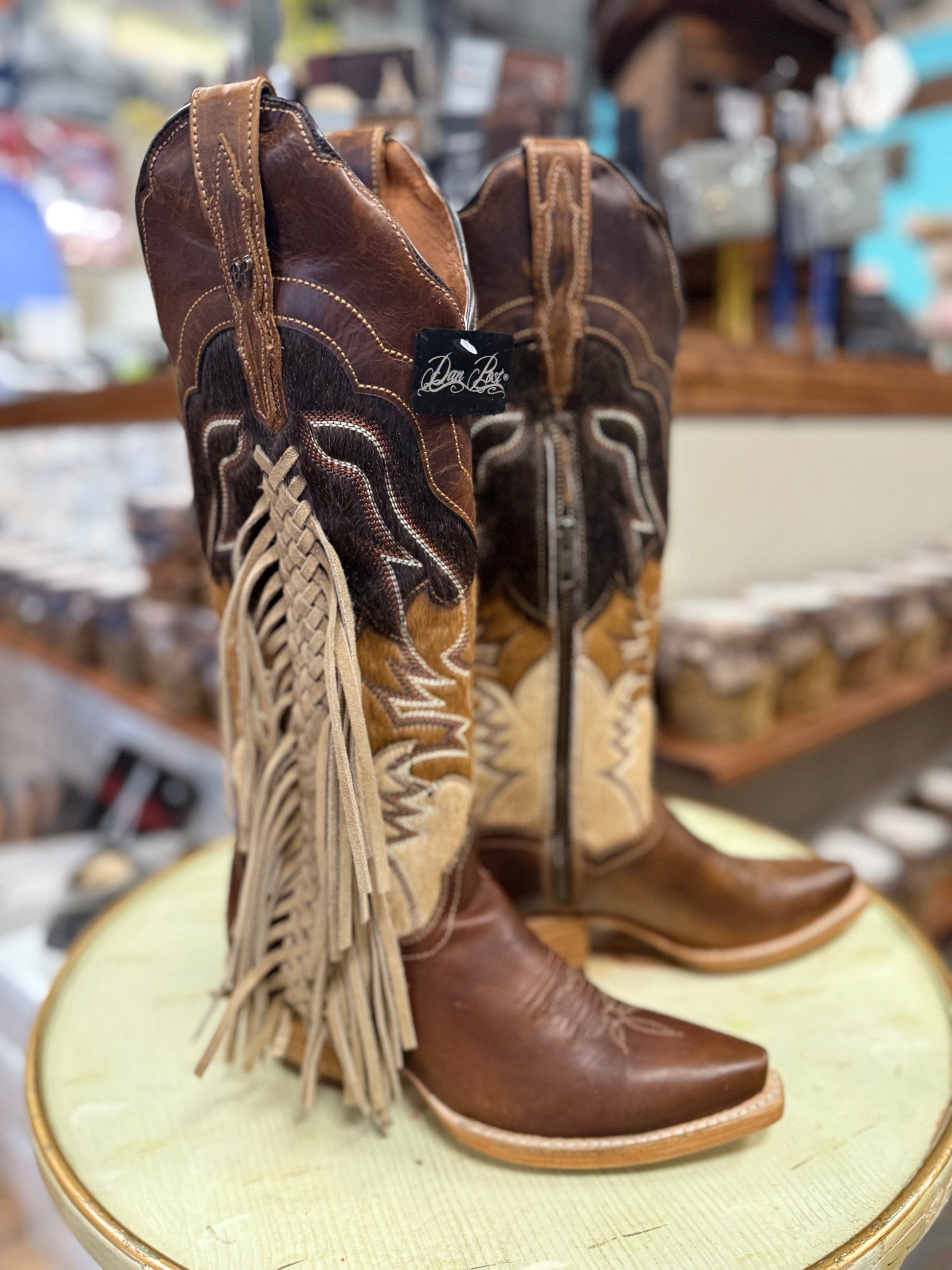 Pair of brown cowboy boots with fringes on a round surface in a store setting.