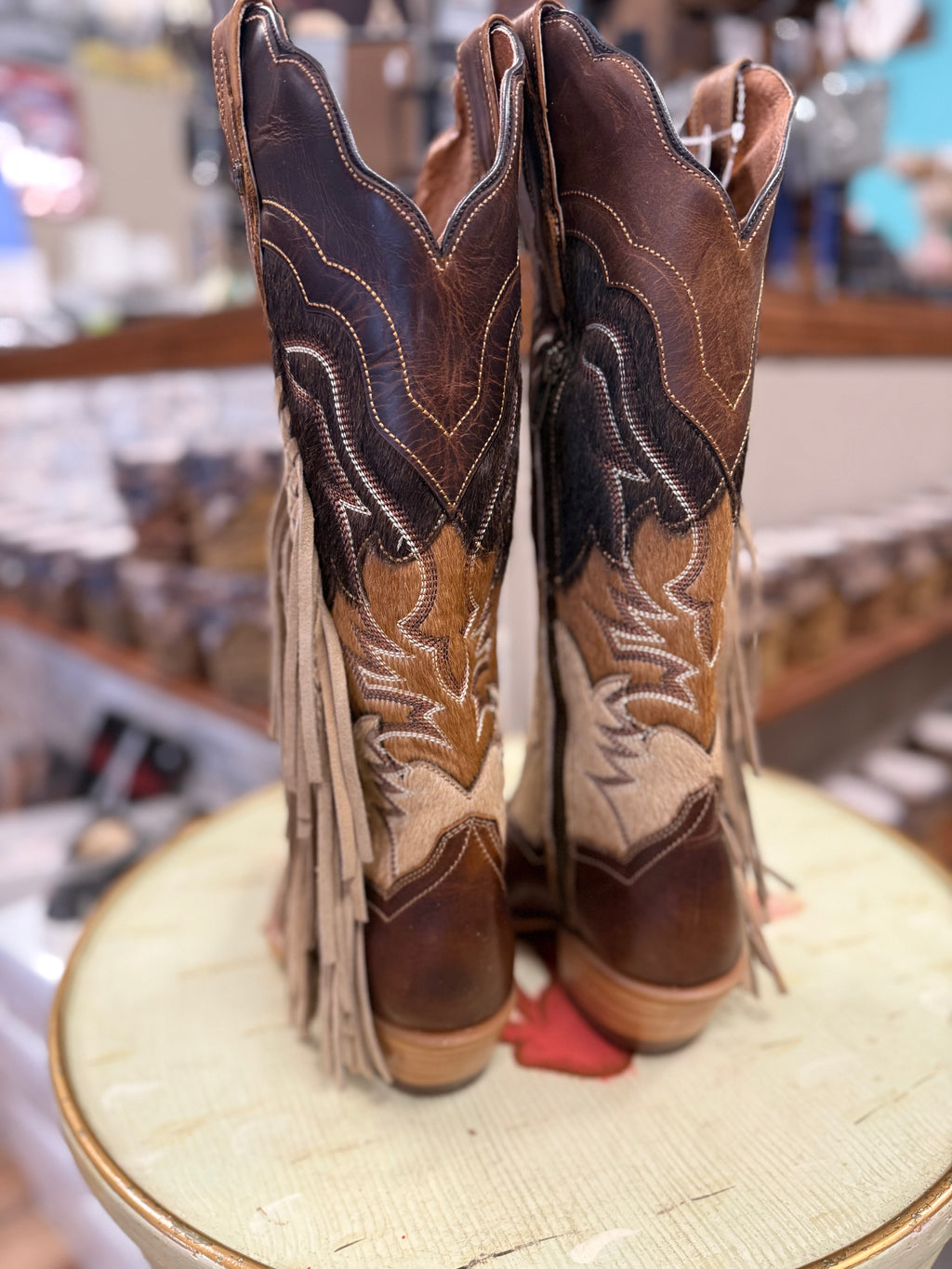 Pair of brown cowboy boots with intricate designs and fringes on a display stand.
