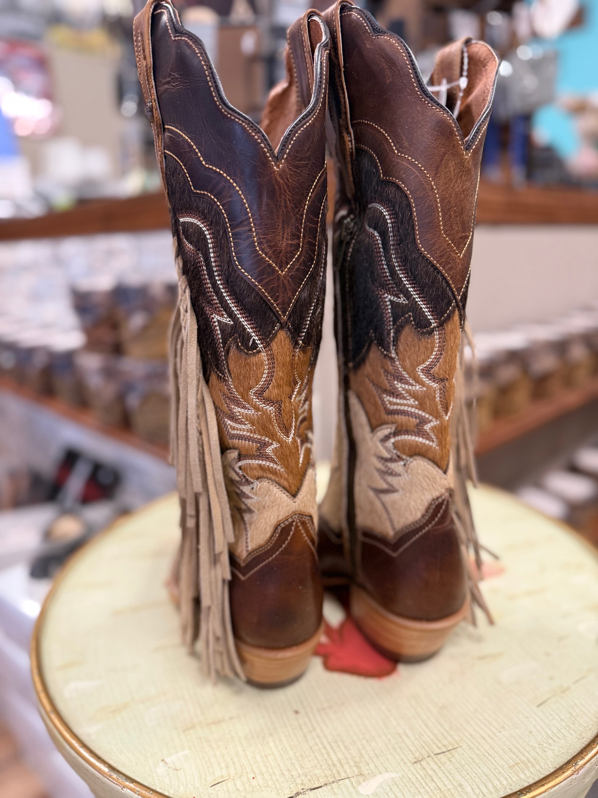Pair of brown cowboy boots with intricate designs and fringes on a display stand.