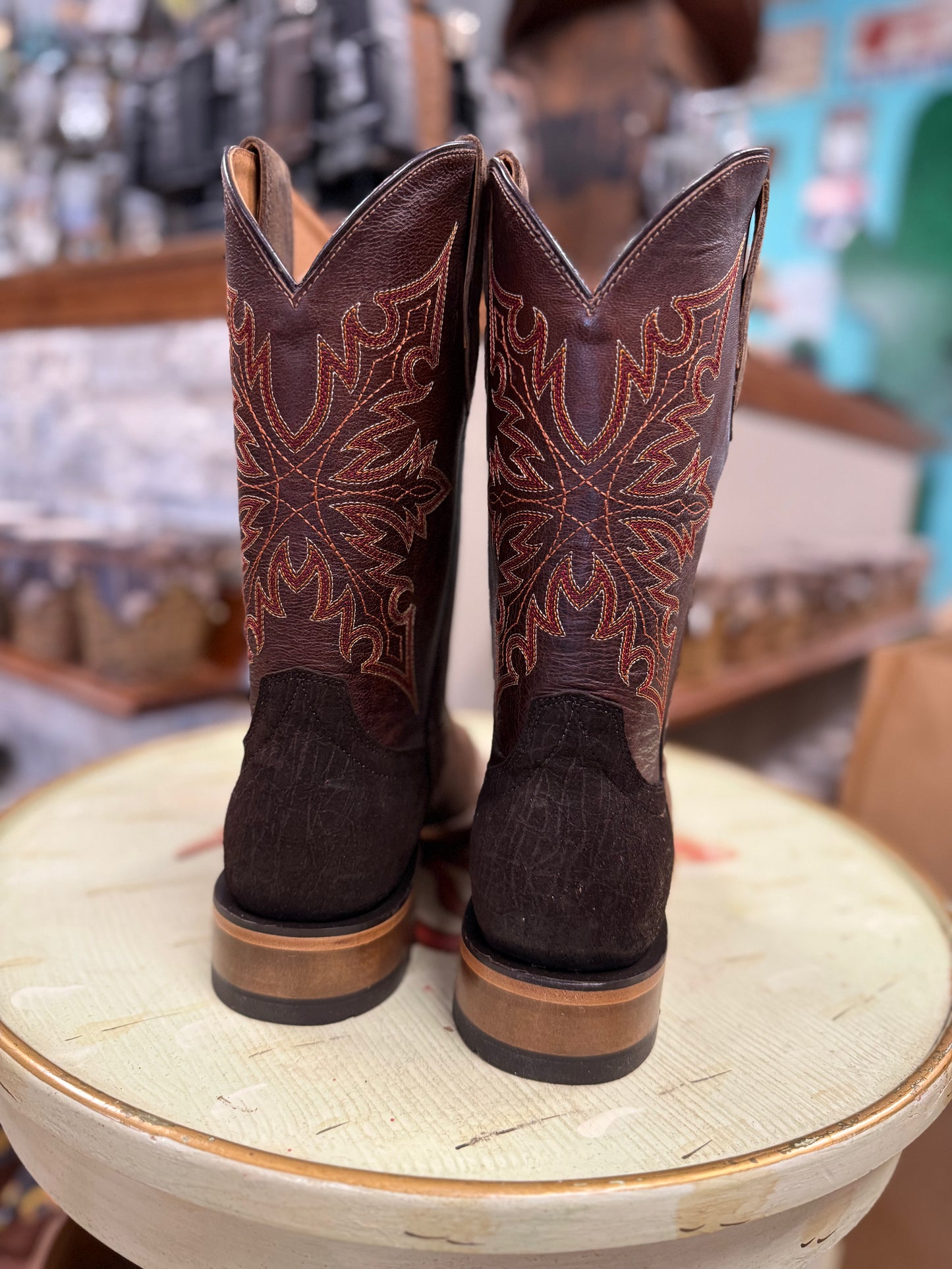 Pair of brown cowboy boots with intricate patterns on a round wooden surface.