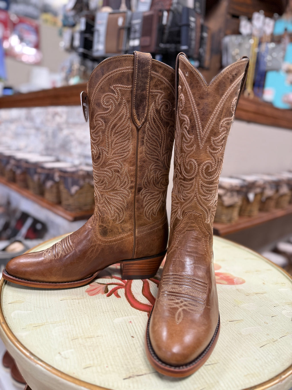 Pair of brown cowboy boots with intricate designs on a stool in a store setting.