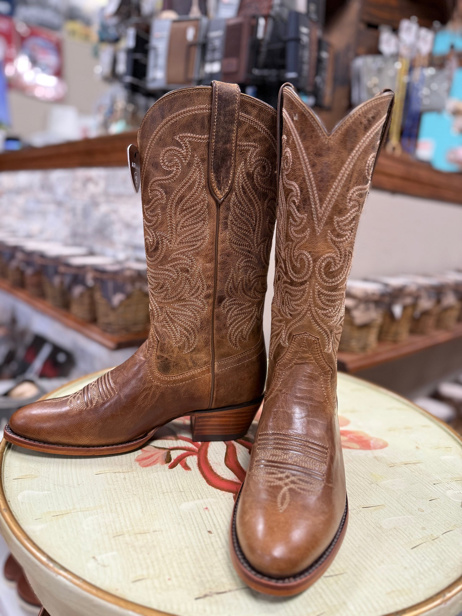 Pair of brown cowboy boots with intricate designs on a stool in a store setting.