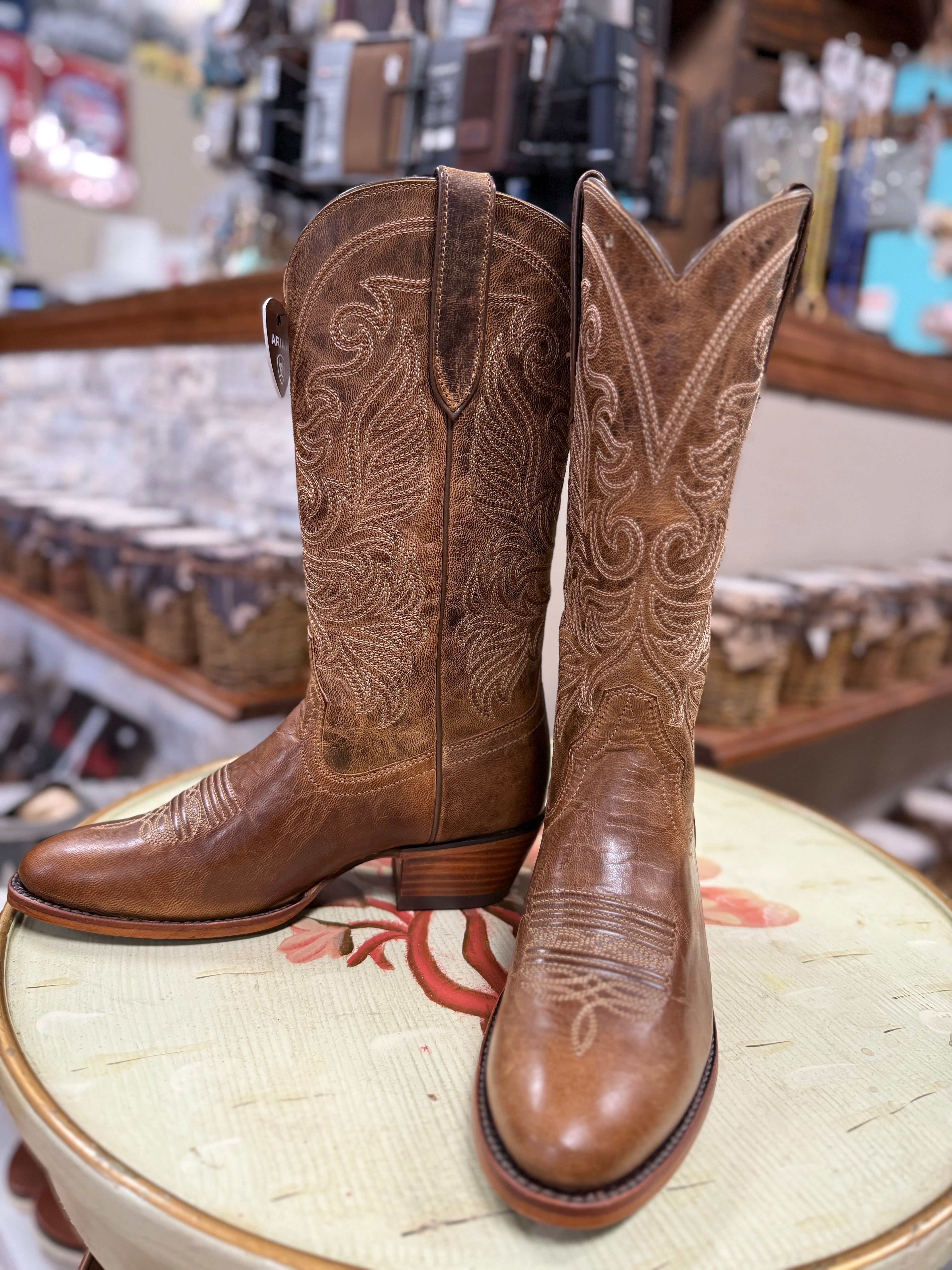 Pair of brown cowboy boots with intricate designs on a stool in a store setting.