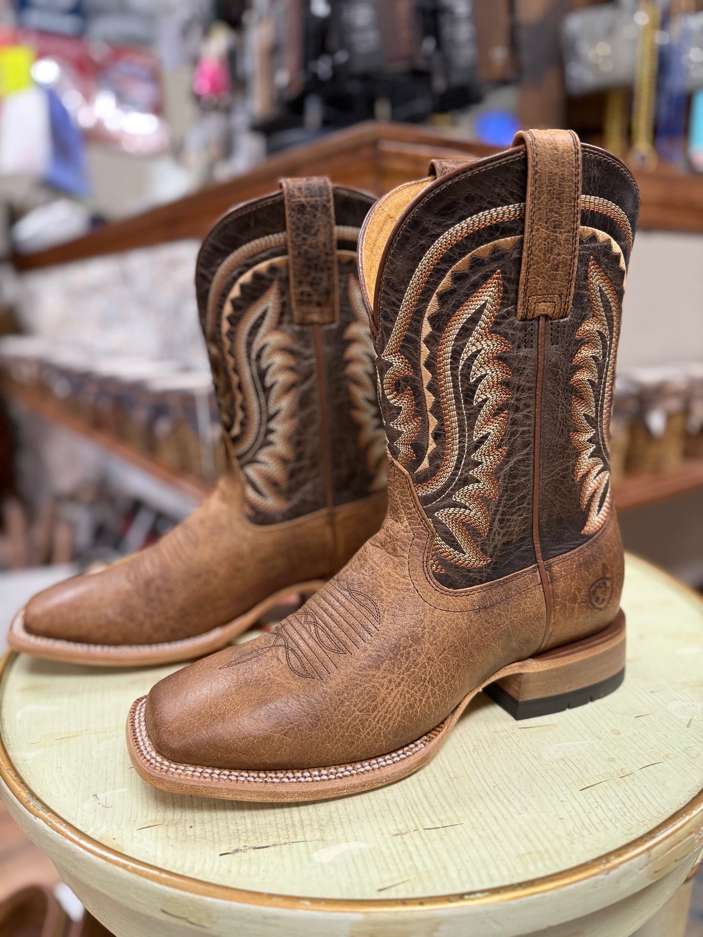 Pair of brown cowboy boots with intricate designs on a round surface in a store setting.