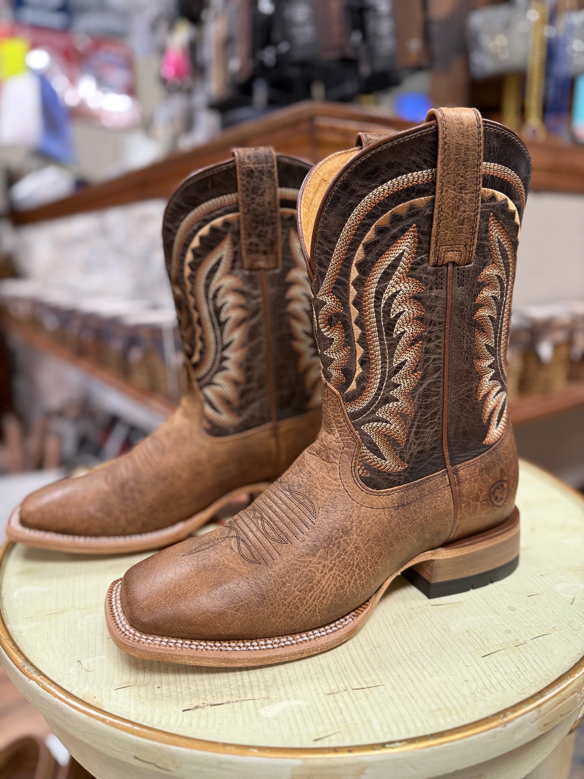 Pair of brown cowboy boots with intricate designs on a round surface in a store setting.