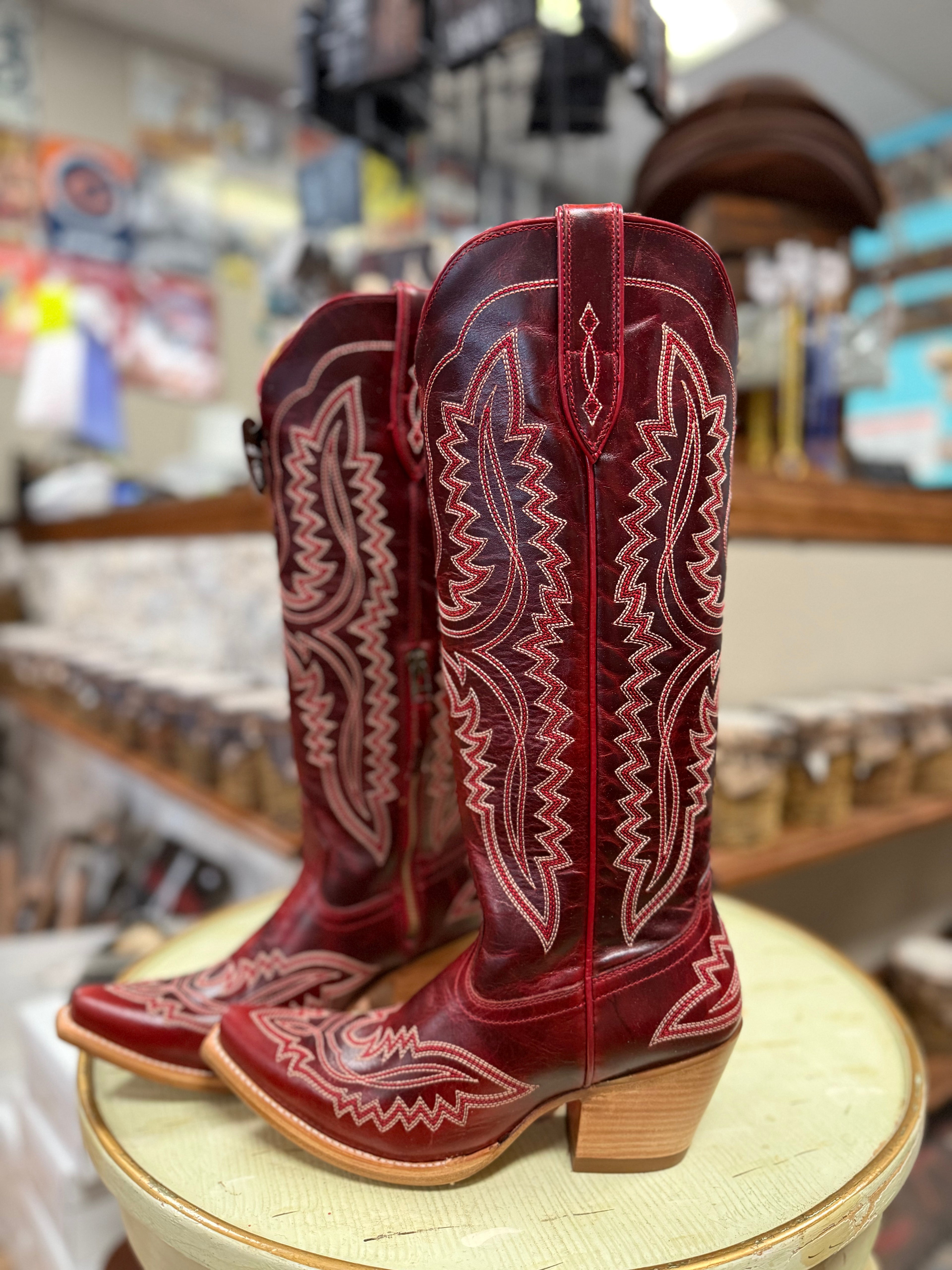 Red cowboy boots with intricate white stitching on a wooden stool in a store setting.