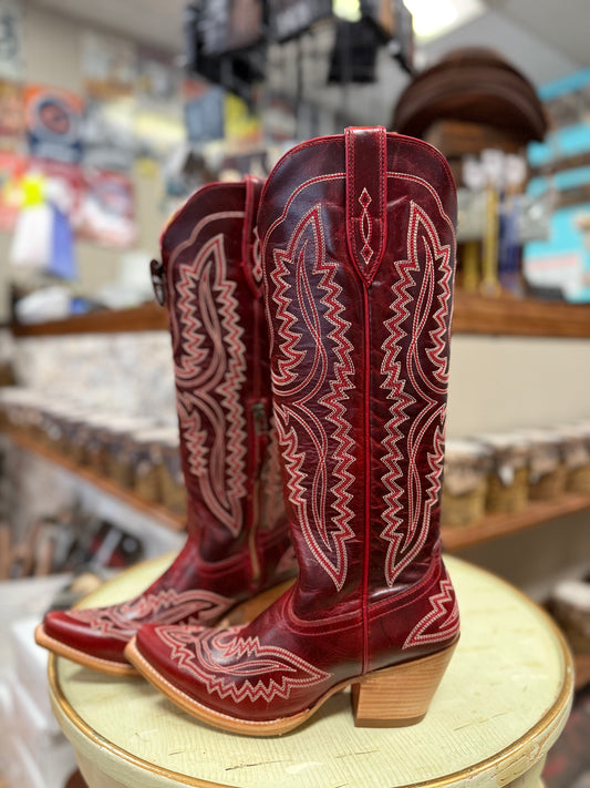 Red cowboy boots with intricate white stitching on a wooden stool in a store setting.
