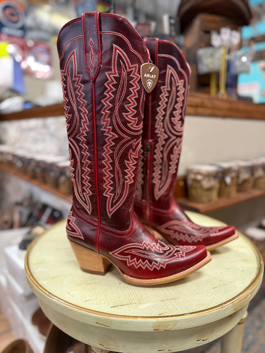 Red cowboy boots with white embroidery on a round wooden stool in a store setting.