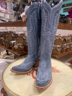 A pair of blue denim western boots with detailed stitching, displayed on a table with a country store background.
