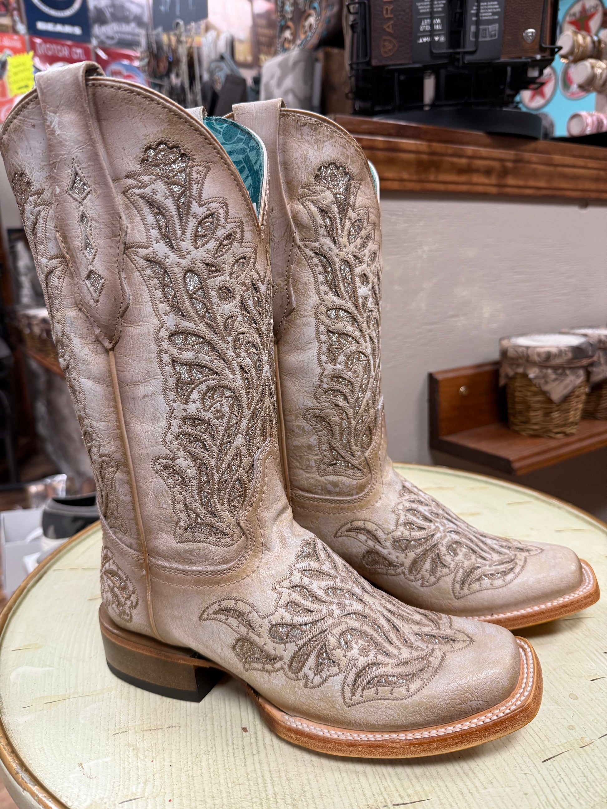 Pair of beige cowboy boots with intricate patterns on a round surface in a store setting.