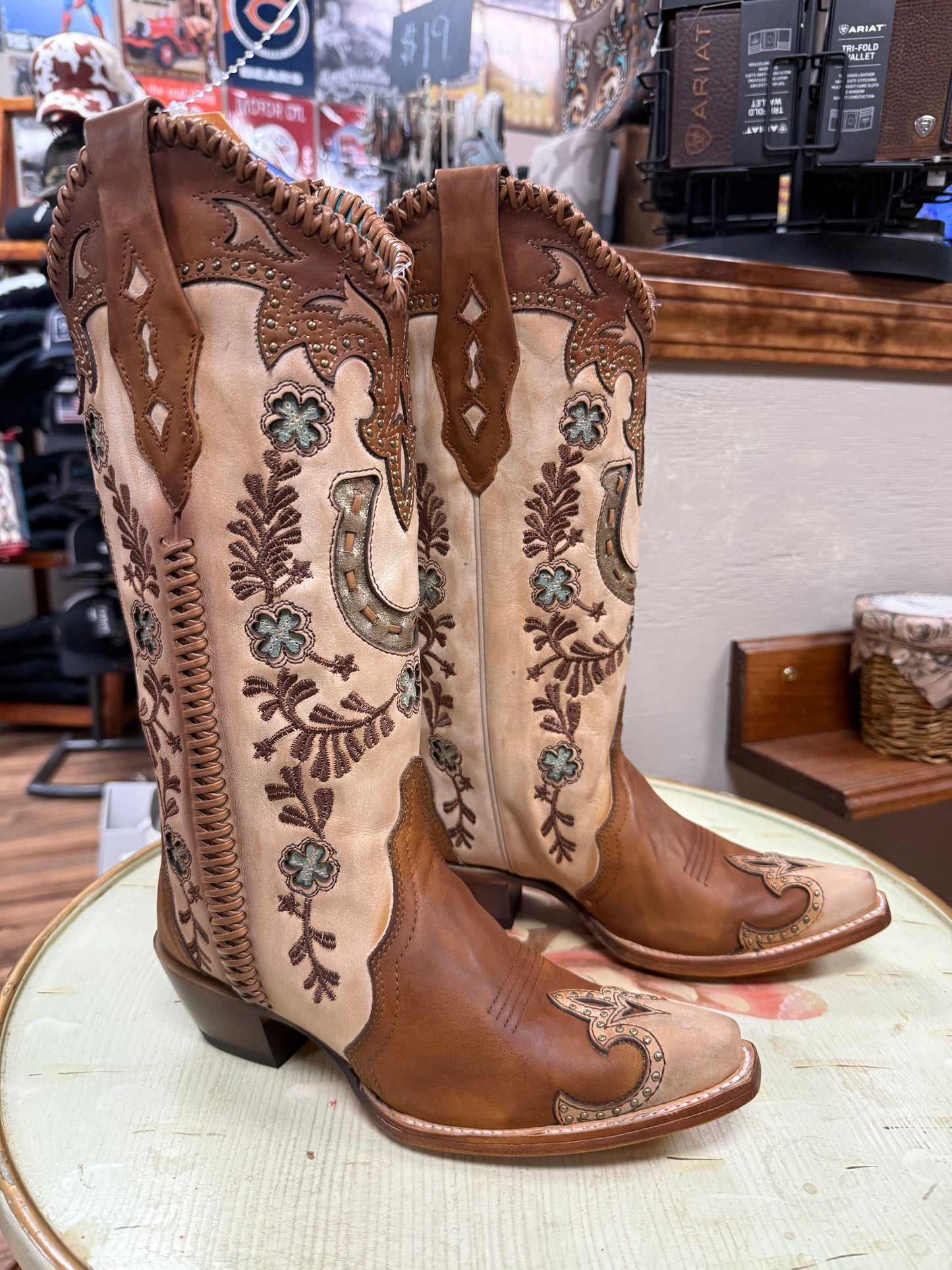 Pair of brown and beige embroidered cowboy boots on a round surface with a store interior background.